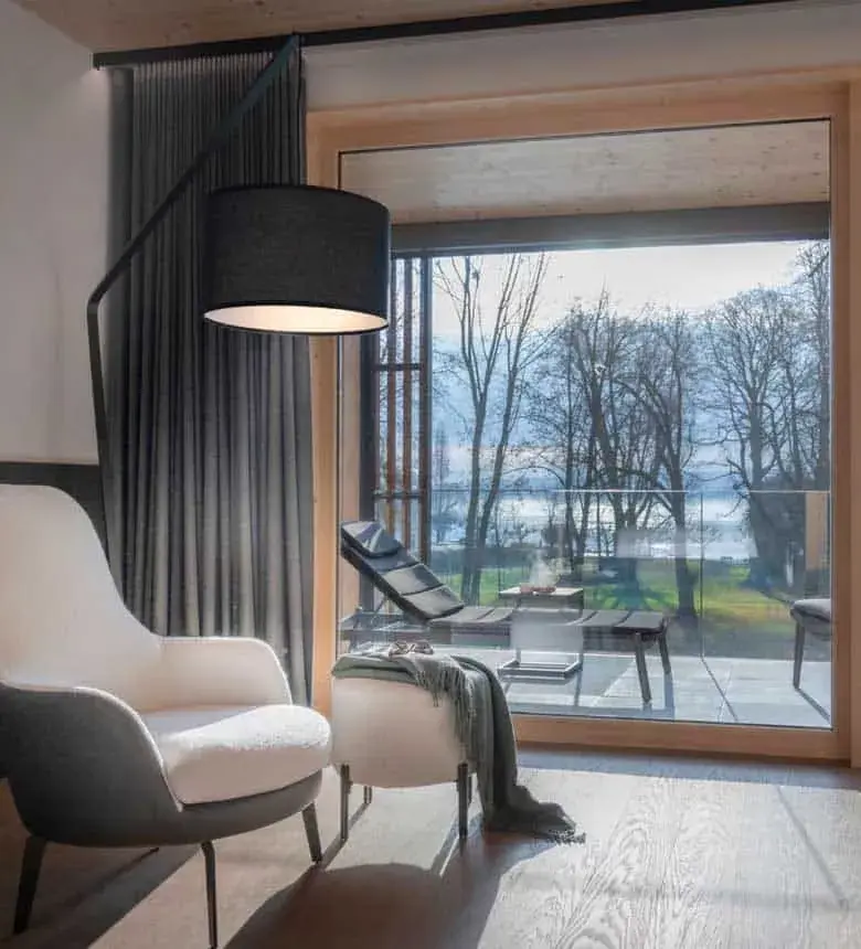 Modern hotel suite at Buff Medical Resort with cosy armchair, floor lamp, and large window overlooking a scenic terrace and lake view.