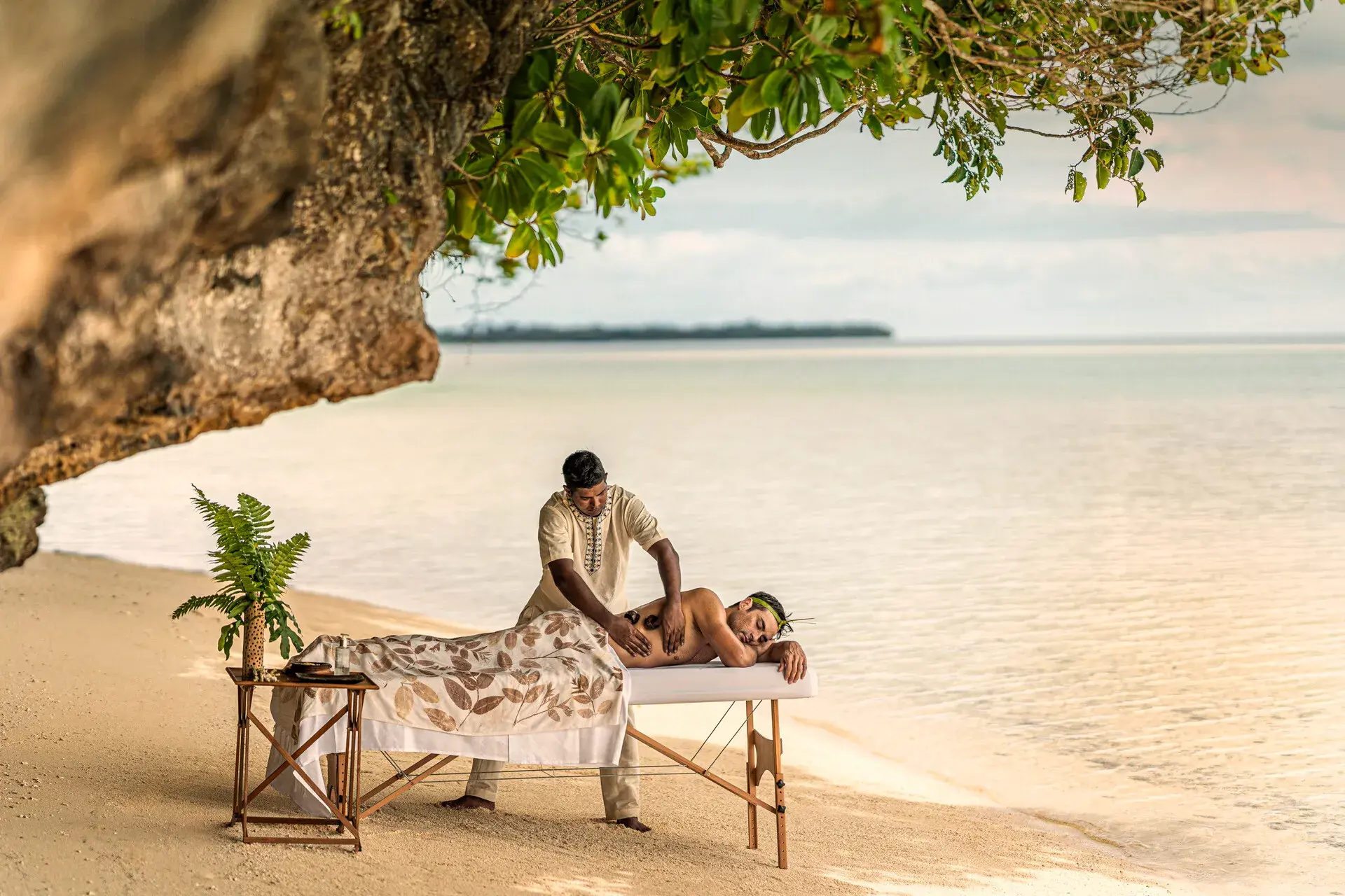 A guest of Four Seasons Explorer Palau receiving a massage from a practitioner on a secluded beach by the water, with green plants hanging above