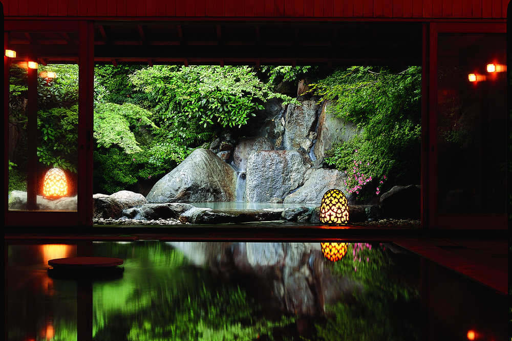 An indoor onsen bath with a view of an outdoor onsen surrounded by rocks through sliding doors