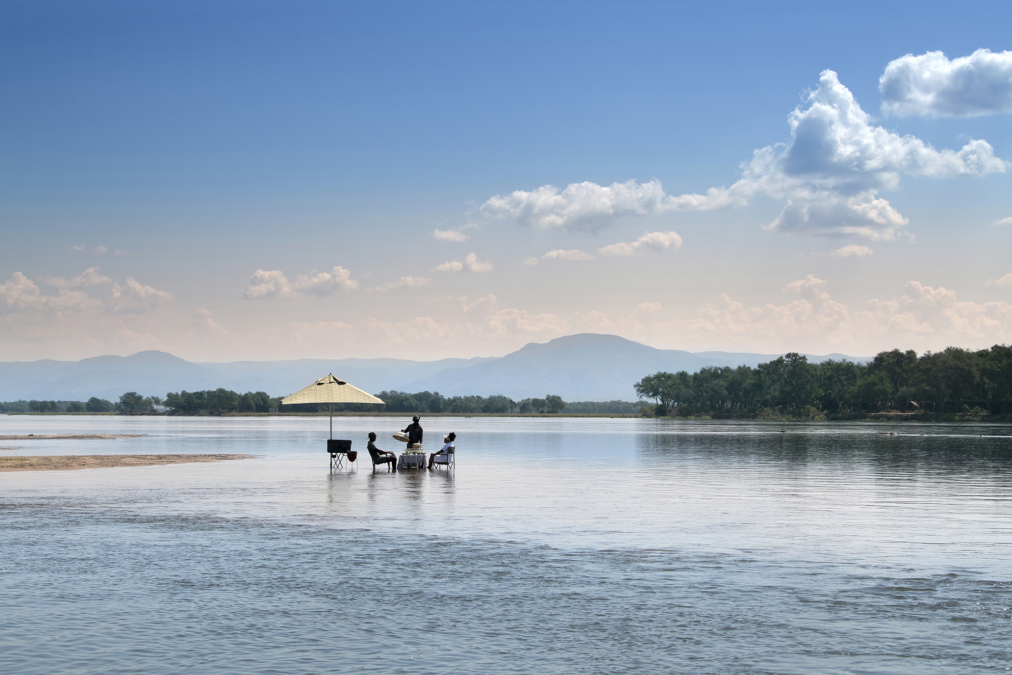 A couple having dinner in the Zambezi river with a waiter and umbrella backed by the mountains under a clear sky