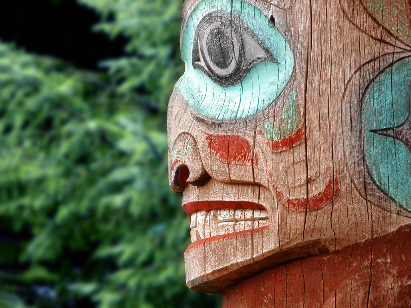 Close-up of a carved wooden totem pole with intricate details, featuring painted eyes and mouth in black, blue, and red against a blurred green background.