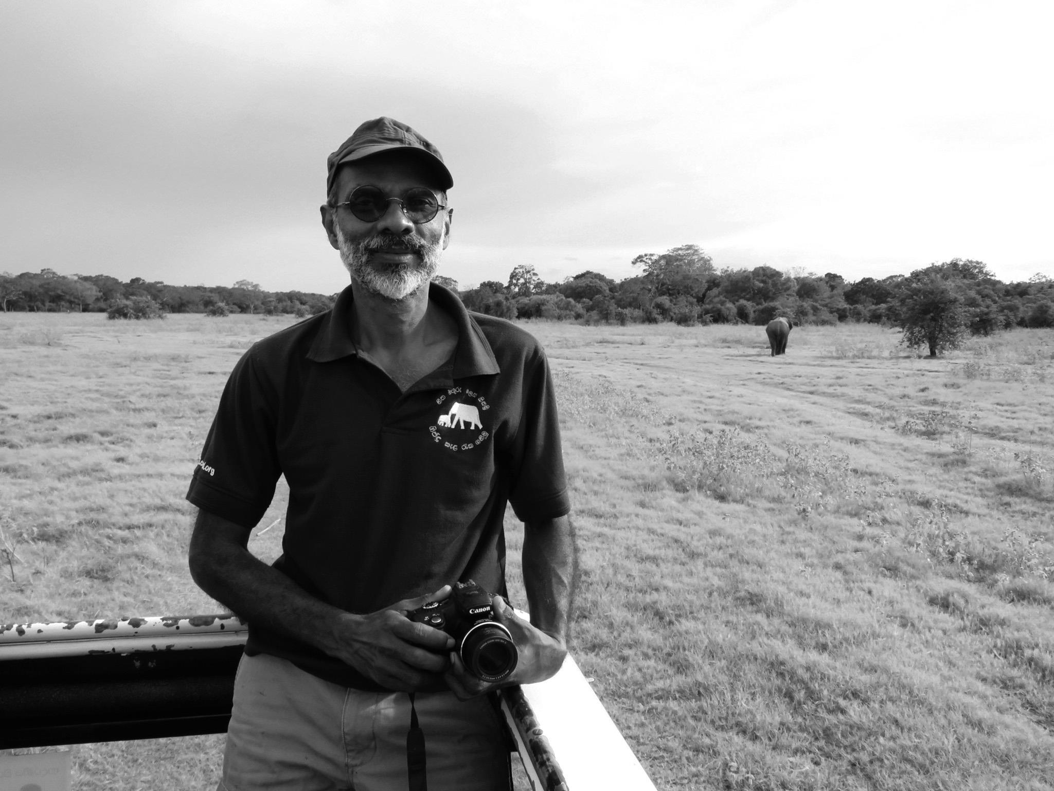 Black and white portrait of Pruthu Fernando holding a camera with elephant in the background
