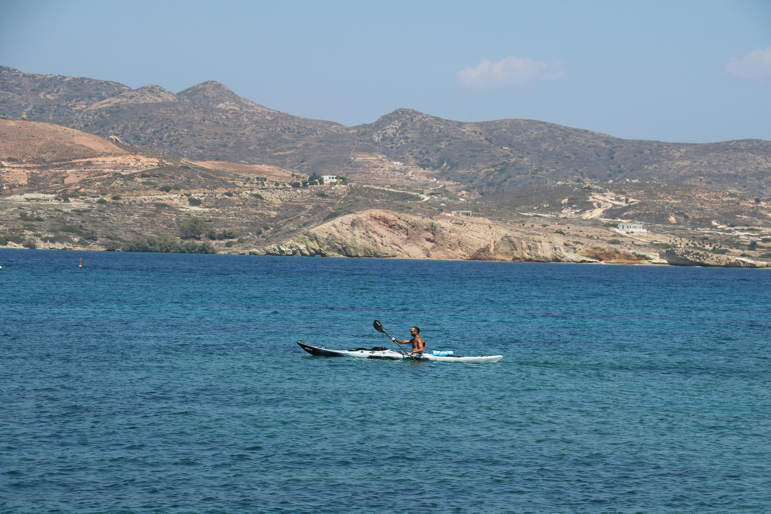 Man paddling a blue sea kayak on calm waters with a rugged and hilly coastline behind 