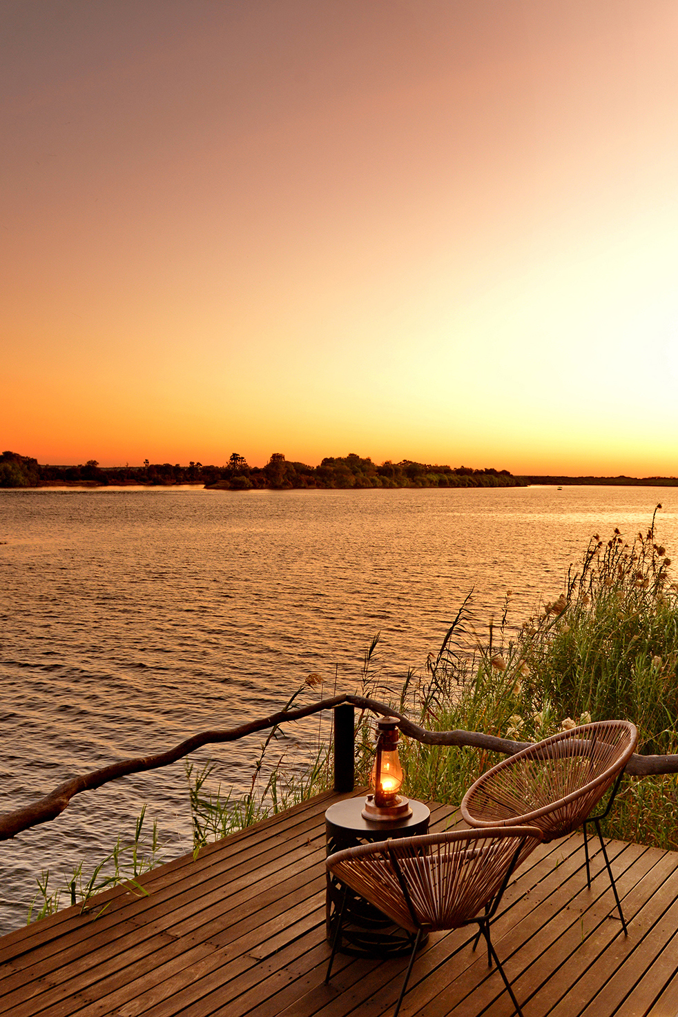 Serene view from Mukwa River Lodge’s wooden deck overlooking a calm river at sunset, with two wicker chairs and a lantern-lit table.