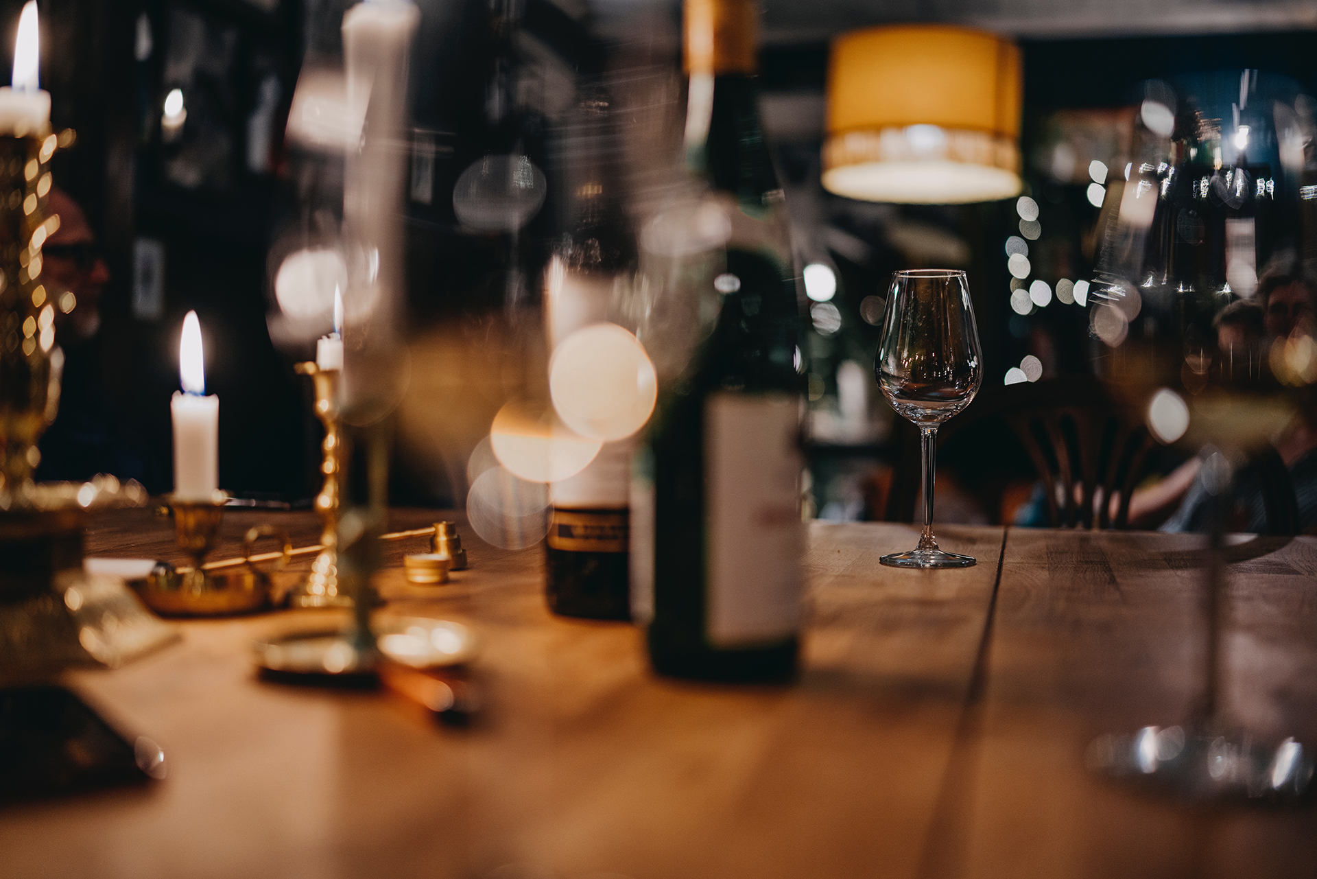 An in focus empty wine glass on a wooden table surrounded by out of focus wine bottle and glasses