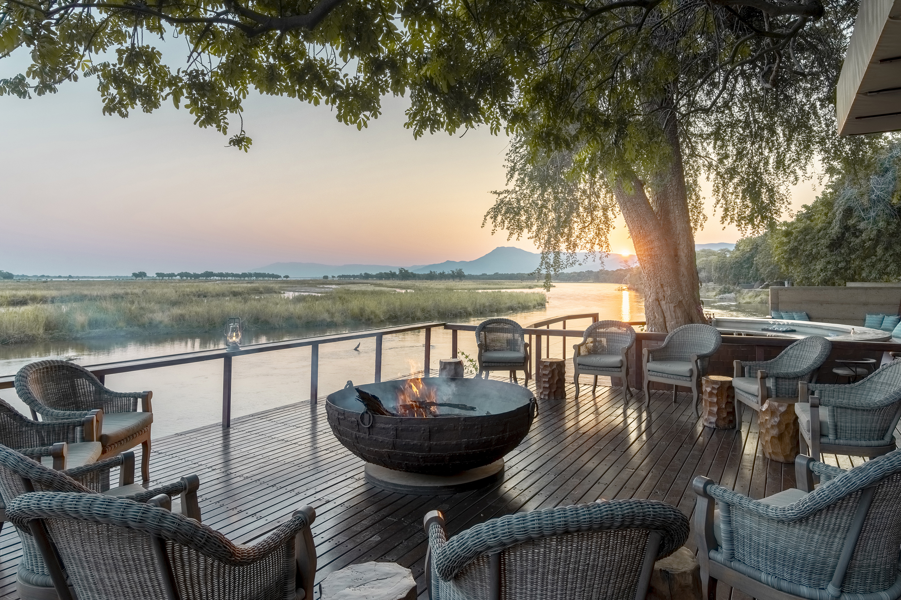 Outdoor seating area with wicker chairs and a fire pit on a wooden deck overlooking the Zambezi River at sunset, surrounded by trees and distant mountains at Sausage Tree Camp in Zambia.