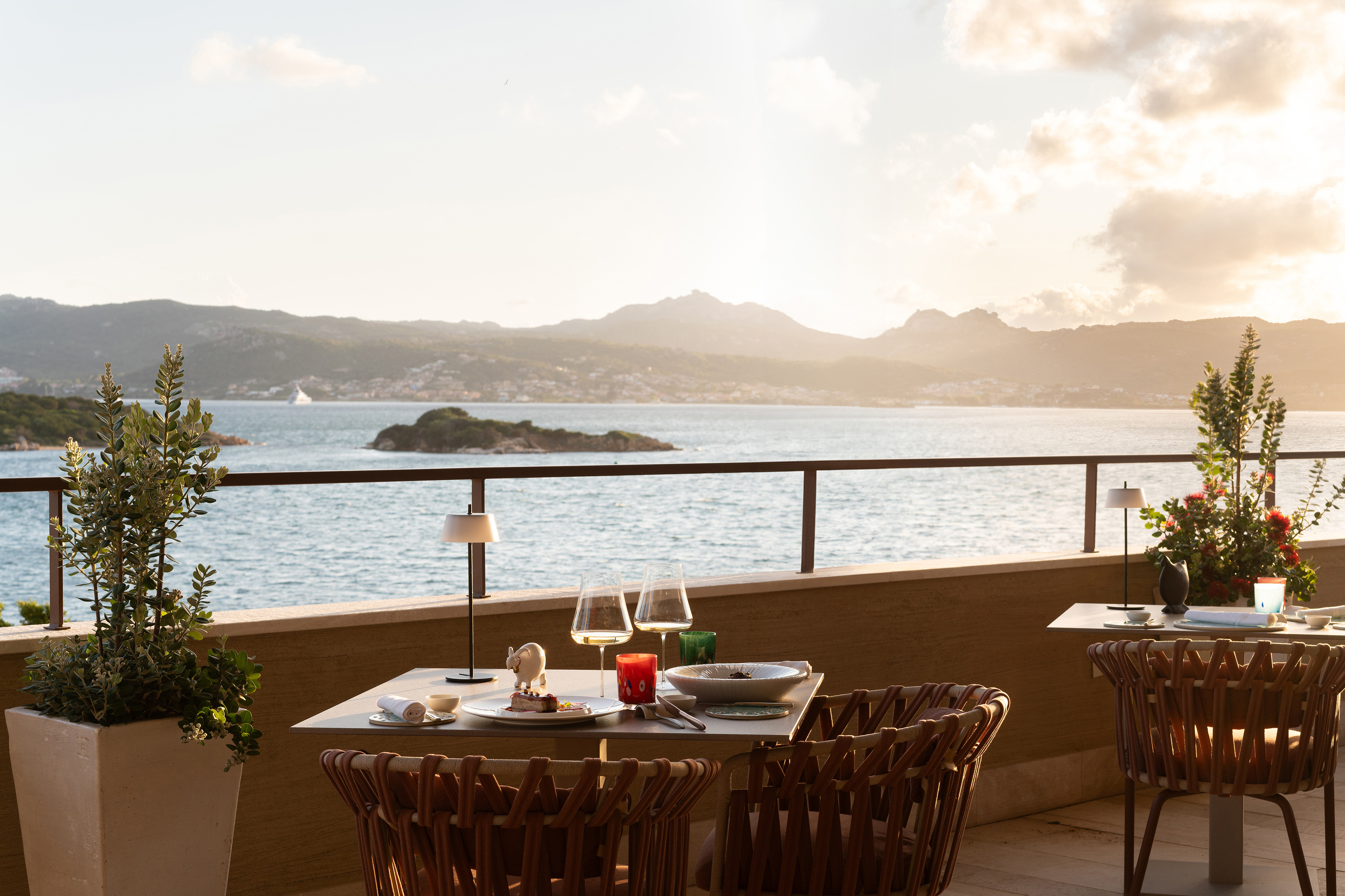 Two tables set up for dinner on an outdoor terrace beside the sea at sunset with a view over a bay