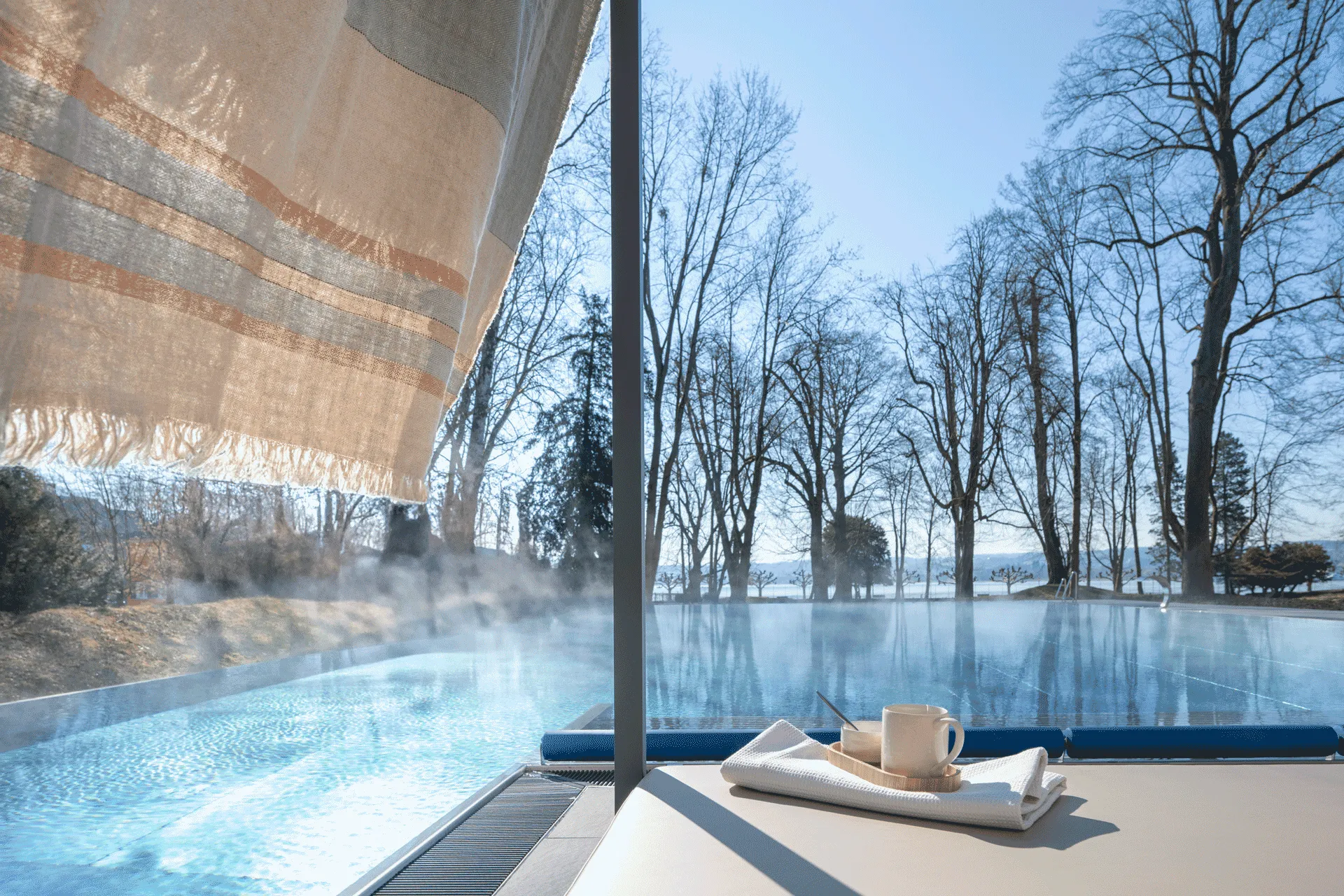 Outdoor infinity pool at Buff Medical Resort with steam rising over still water and tranquil views of trees and the lake beyond.