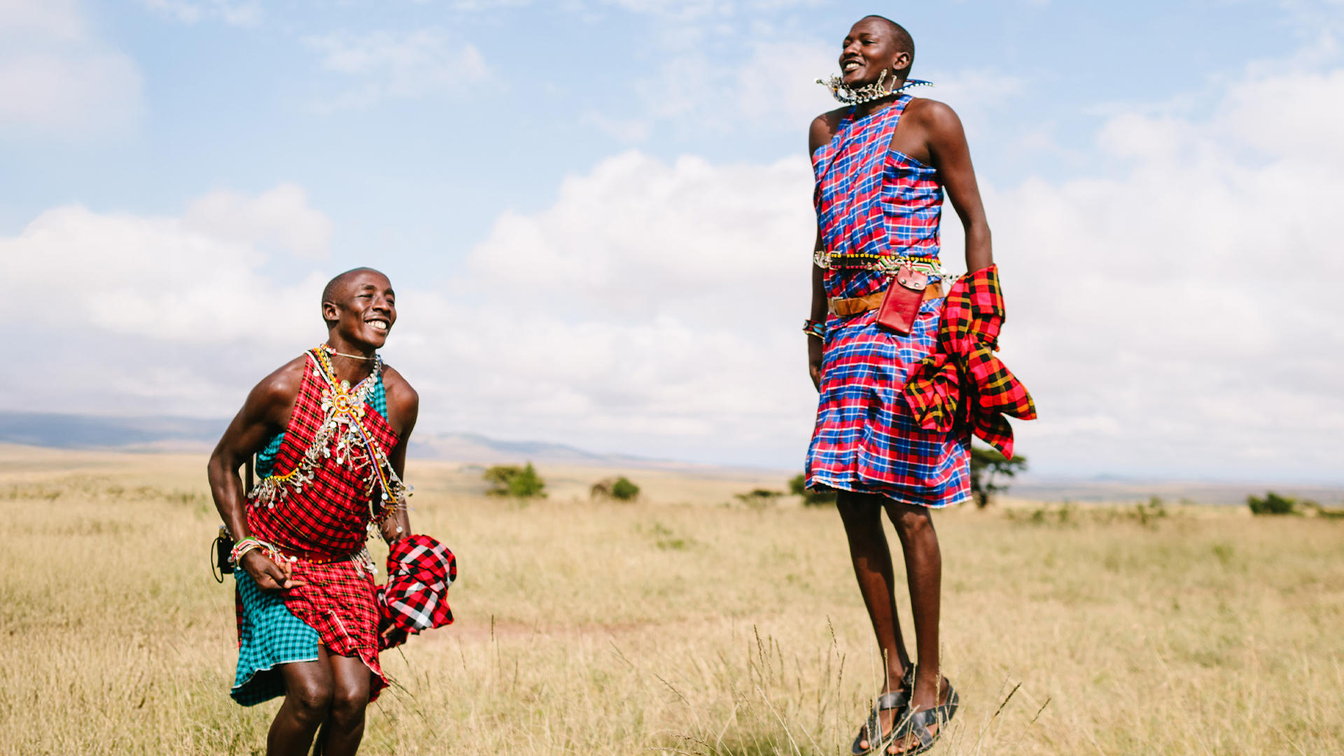 Two Maasai men in colorful traditional attire smile joyfully while jumping high in a grassy field under a blue sky, sharing their culture with families visiting the Kenyan savannah