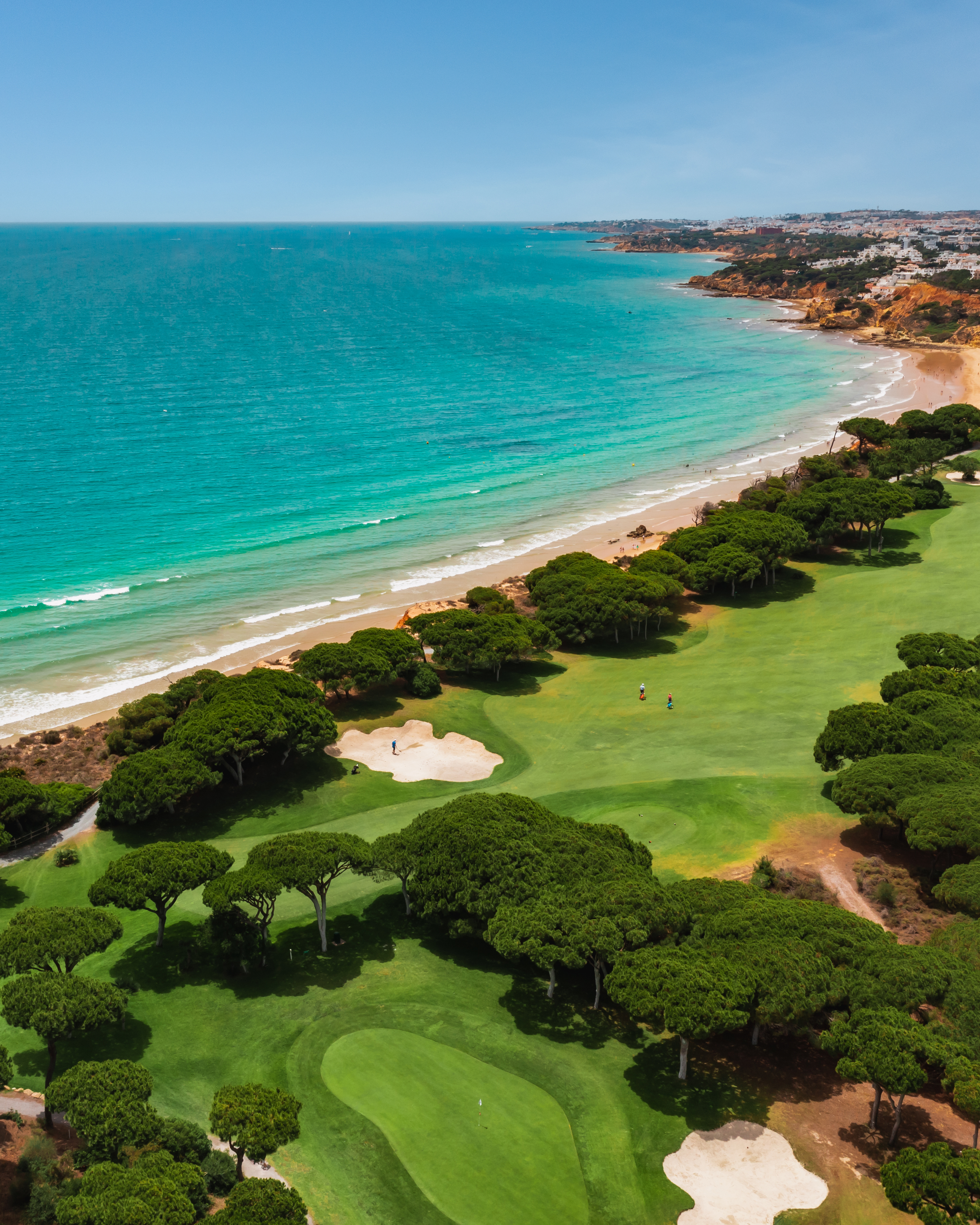 Aerial view of a coastal golf course with sand bunkers, trees, two people playing, and a nearby beach with turquoise water, rocky cliffs, and buildings in the background