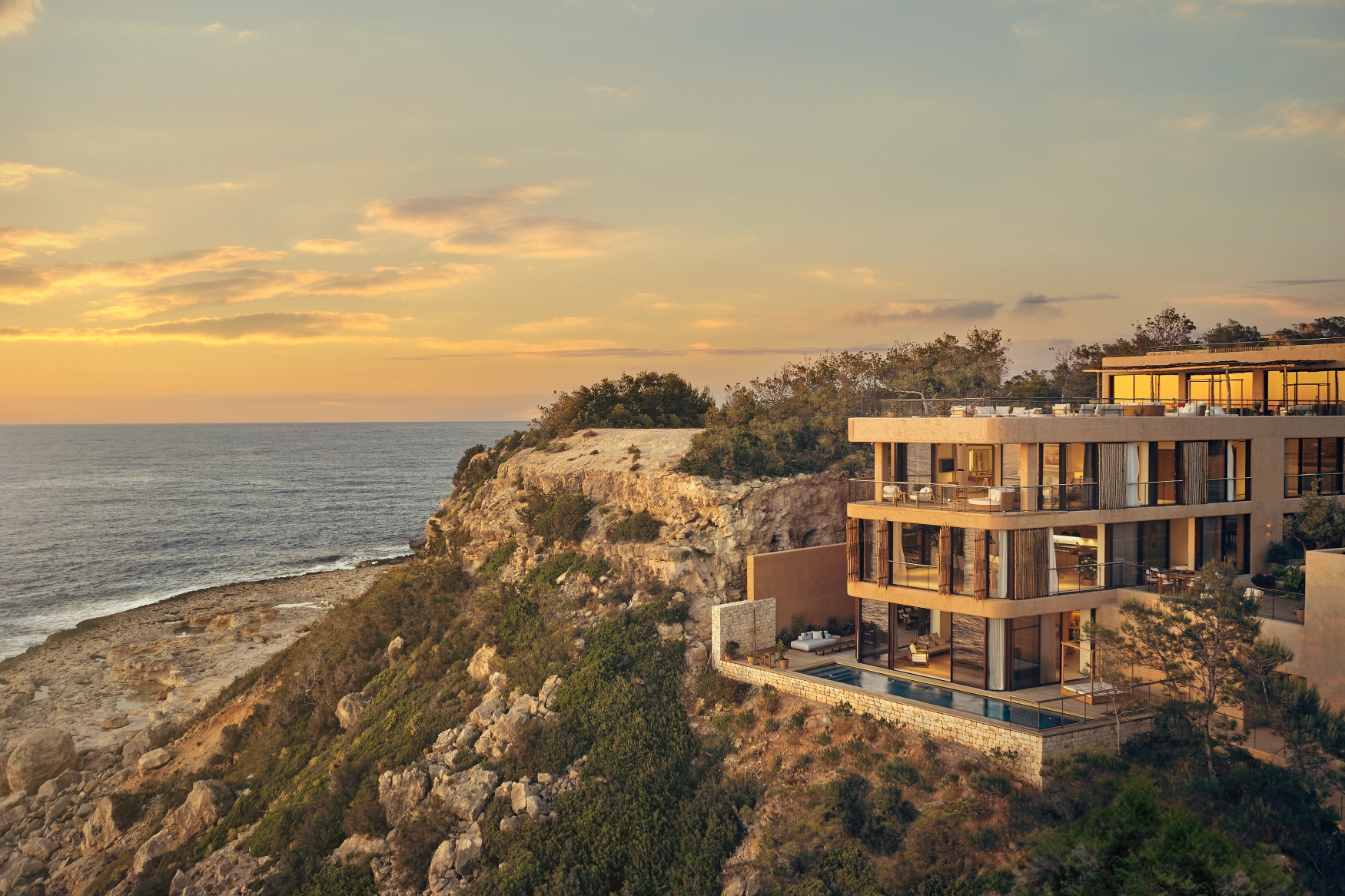 A modern luxury house on a rocky cliff by the ocean at sunset, with large glass windows, terraces, and an infinity pool.