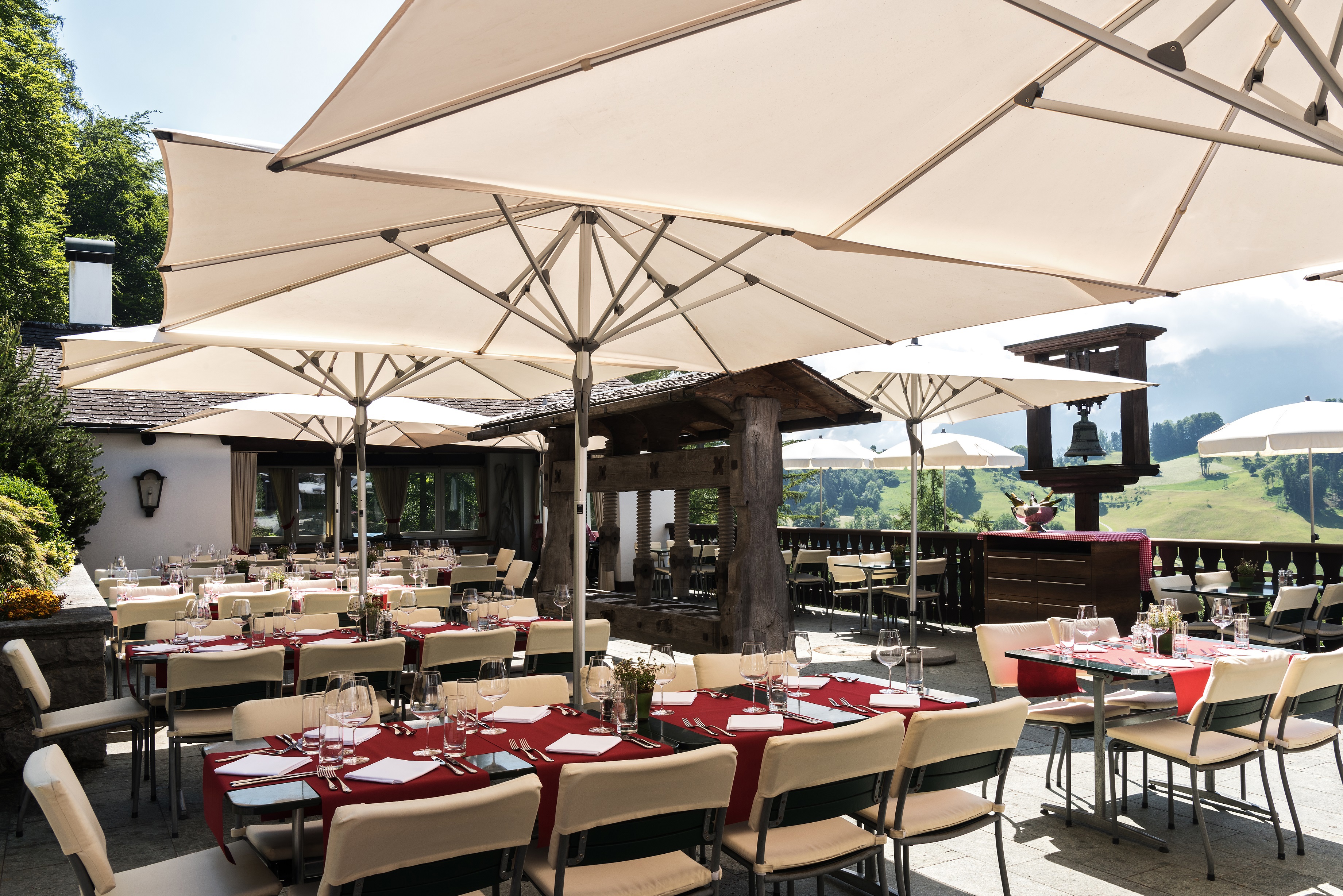 The outdoor terrace of the Taverne featuring large dining tables under white sun umbrellas surrounding period features like an old bell