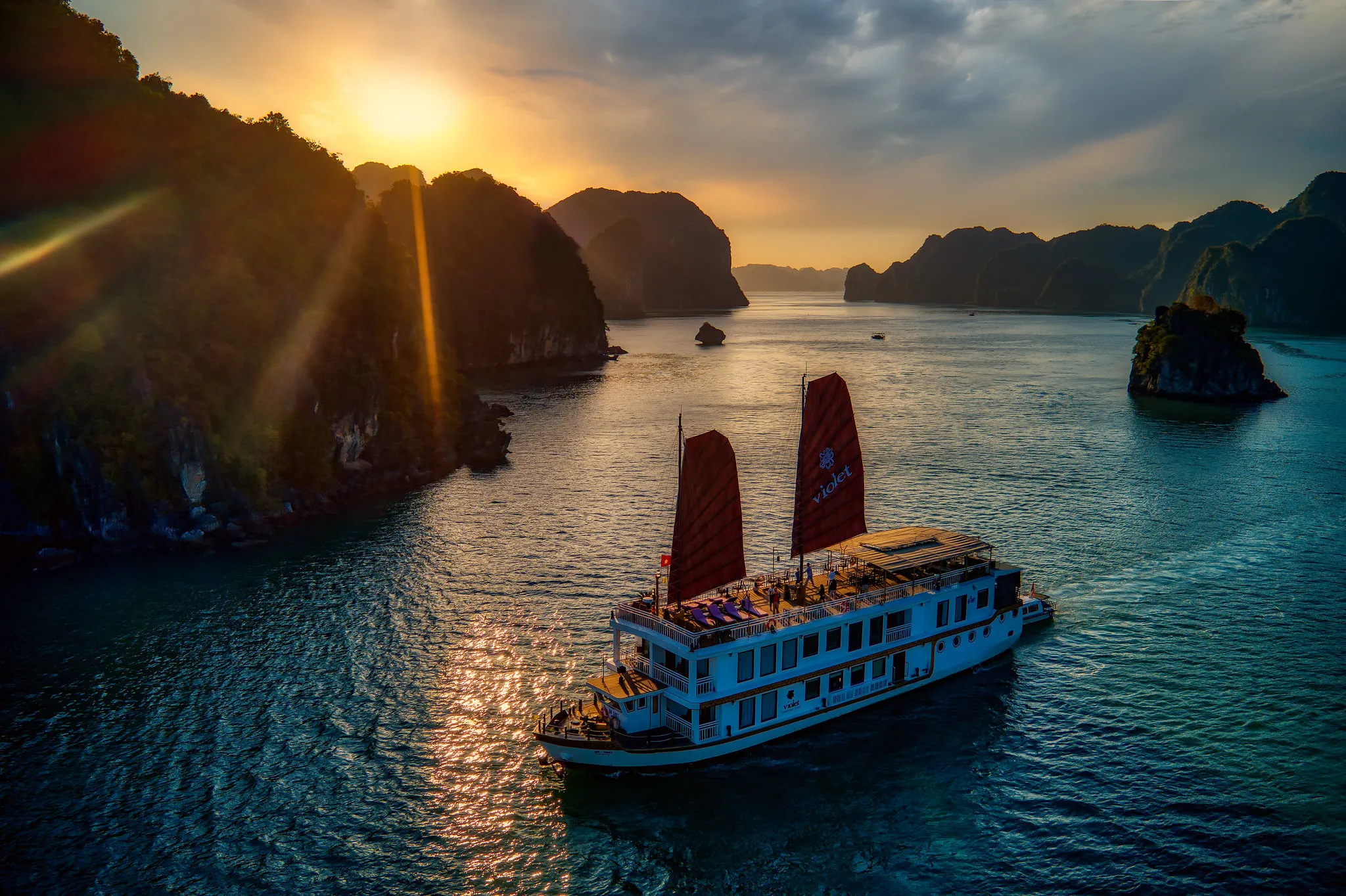 Heritage Line boat with red sails gliding through a tranquil bay at sunset, framed by rocky islands.