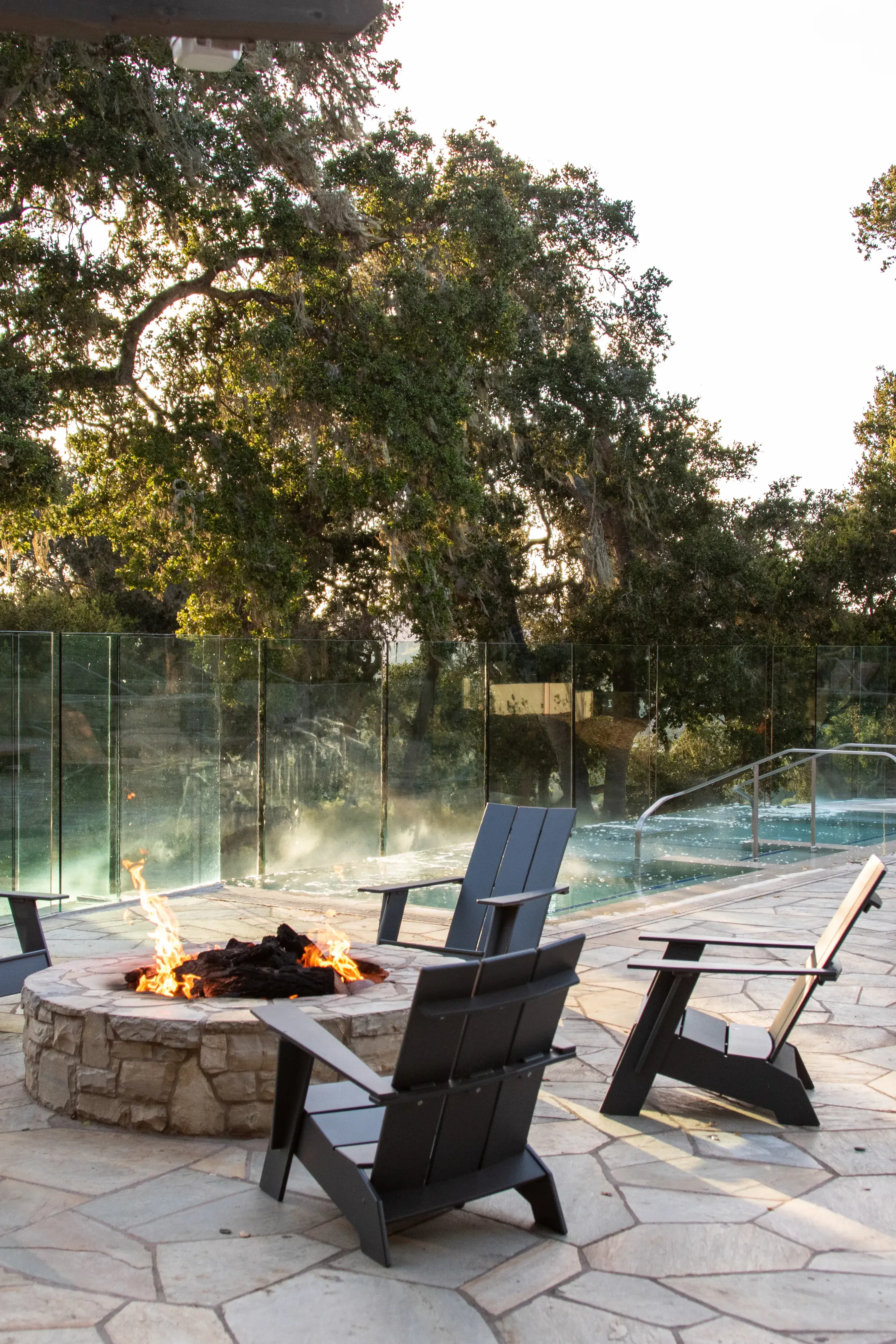 A firepit with wooden chairs on a stone terrace beside the adult pool at Carmel Valley Ranch in California with a view of tall trees