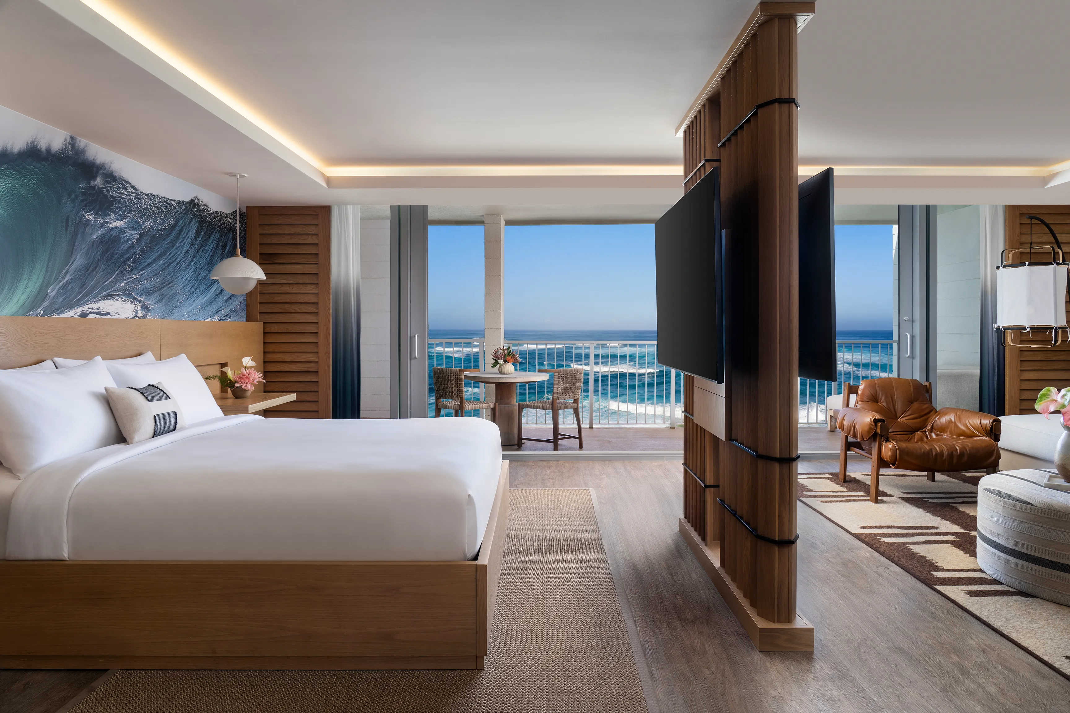 An Ali'i Oceanfront Suite Bedroom at Ritz-Carlton Turtle Bay featuring a tv mounted room divider with a plush bed on one side and a lounge on the other, with large balcony doors overlooking the ocean in the background