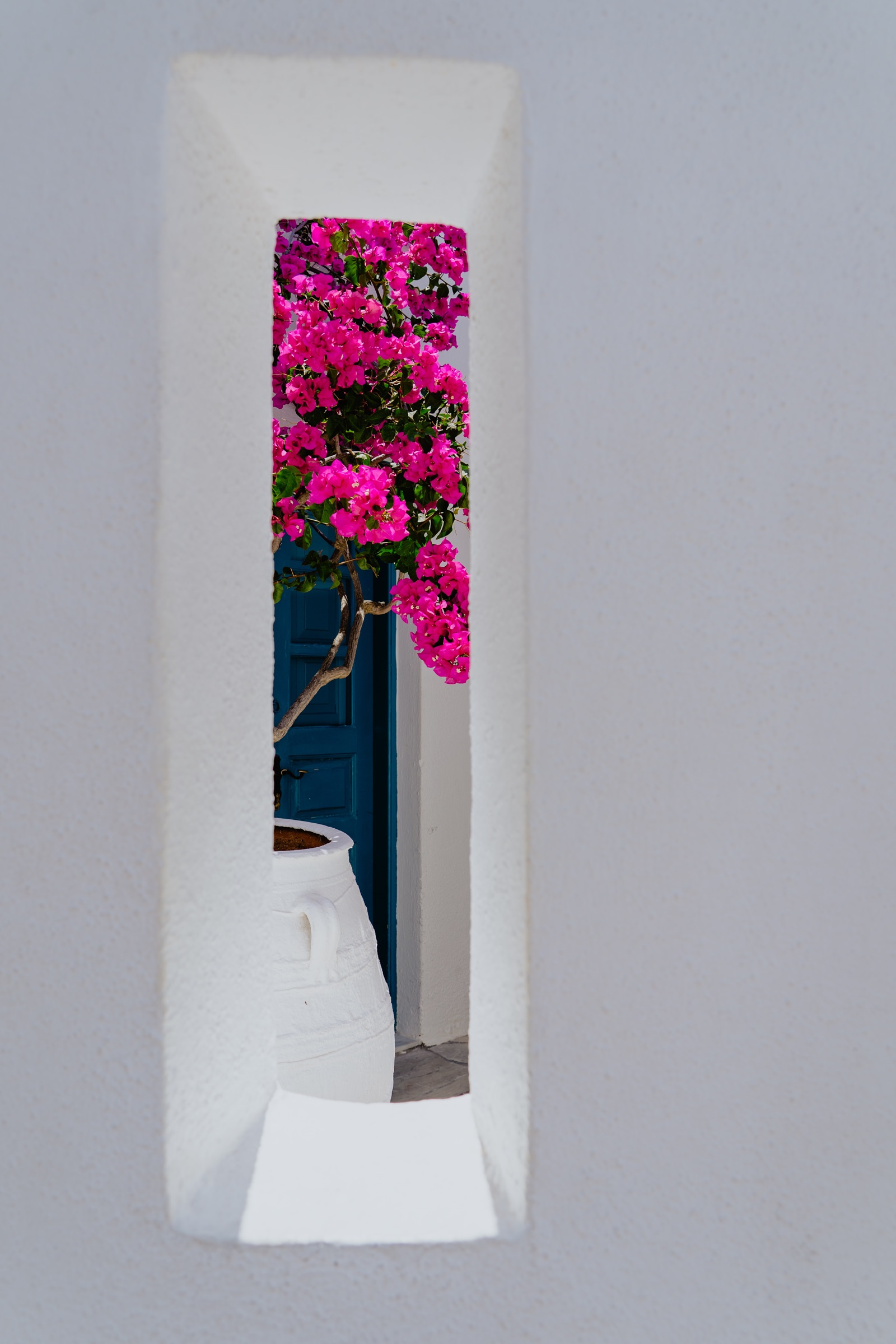 A narrow rectangular window in a whitewashed wall frames a view of pink bougainvillea flowers, a white ceramic pot, and a blue door.