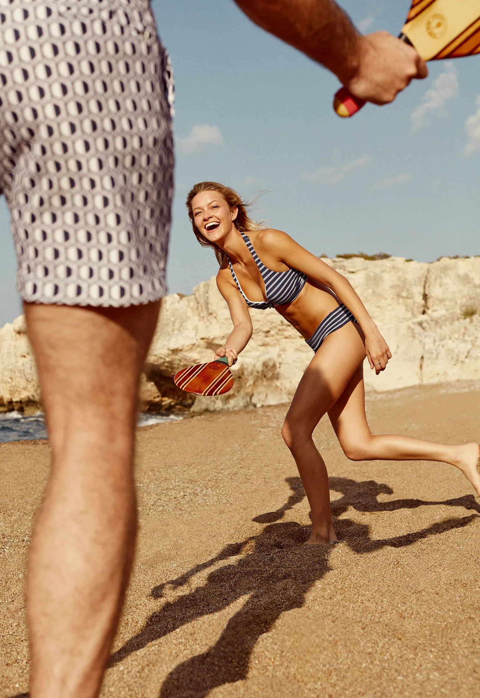 A man and woman in swimwear play padel tennis on a beach with rocks in the background