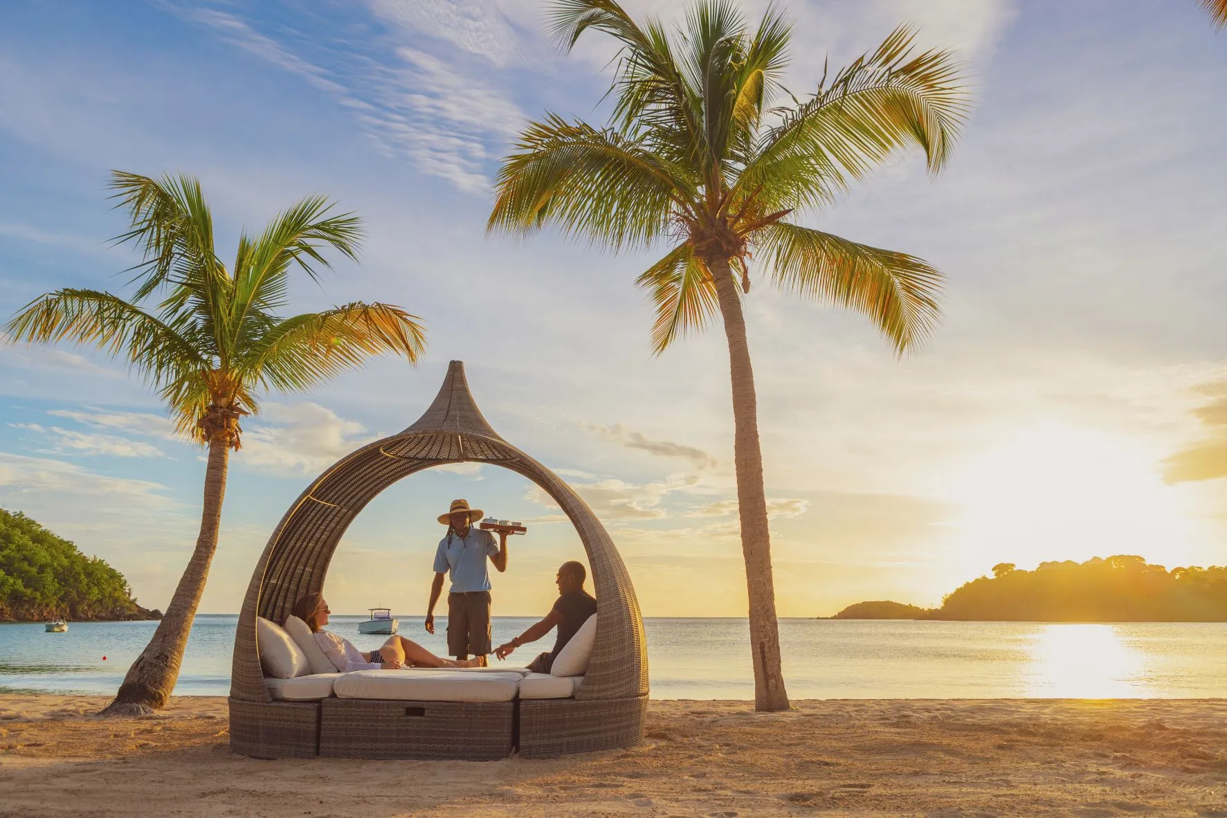 A large sun lounger structure between two palms on the beach at Carlisle Bay with two guests receiving drinks at sunset in front of the ocean