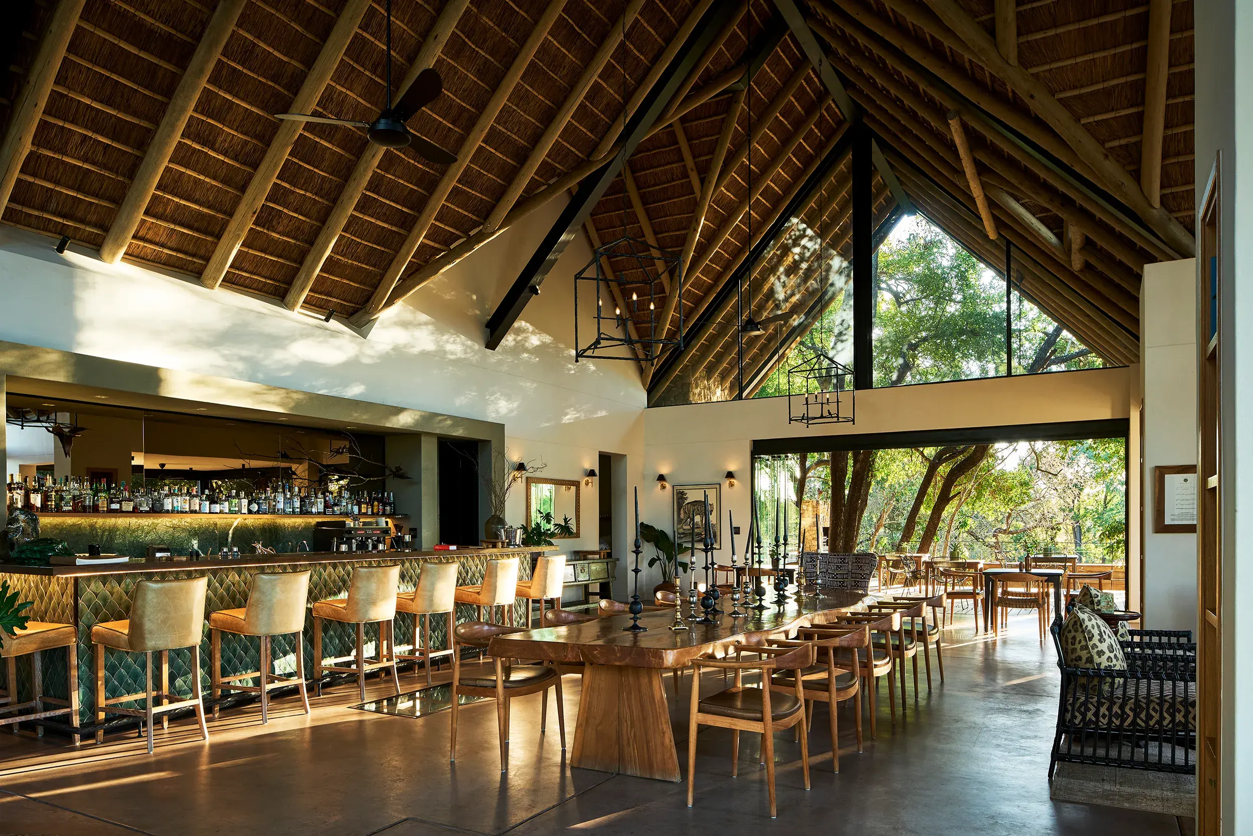 The main dining room at Monwana Game Reserve featuring tall vaulted wooden ceilings above a long wooden table and well stocked bar with a view of the trees outside through a large glass wall