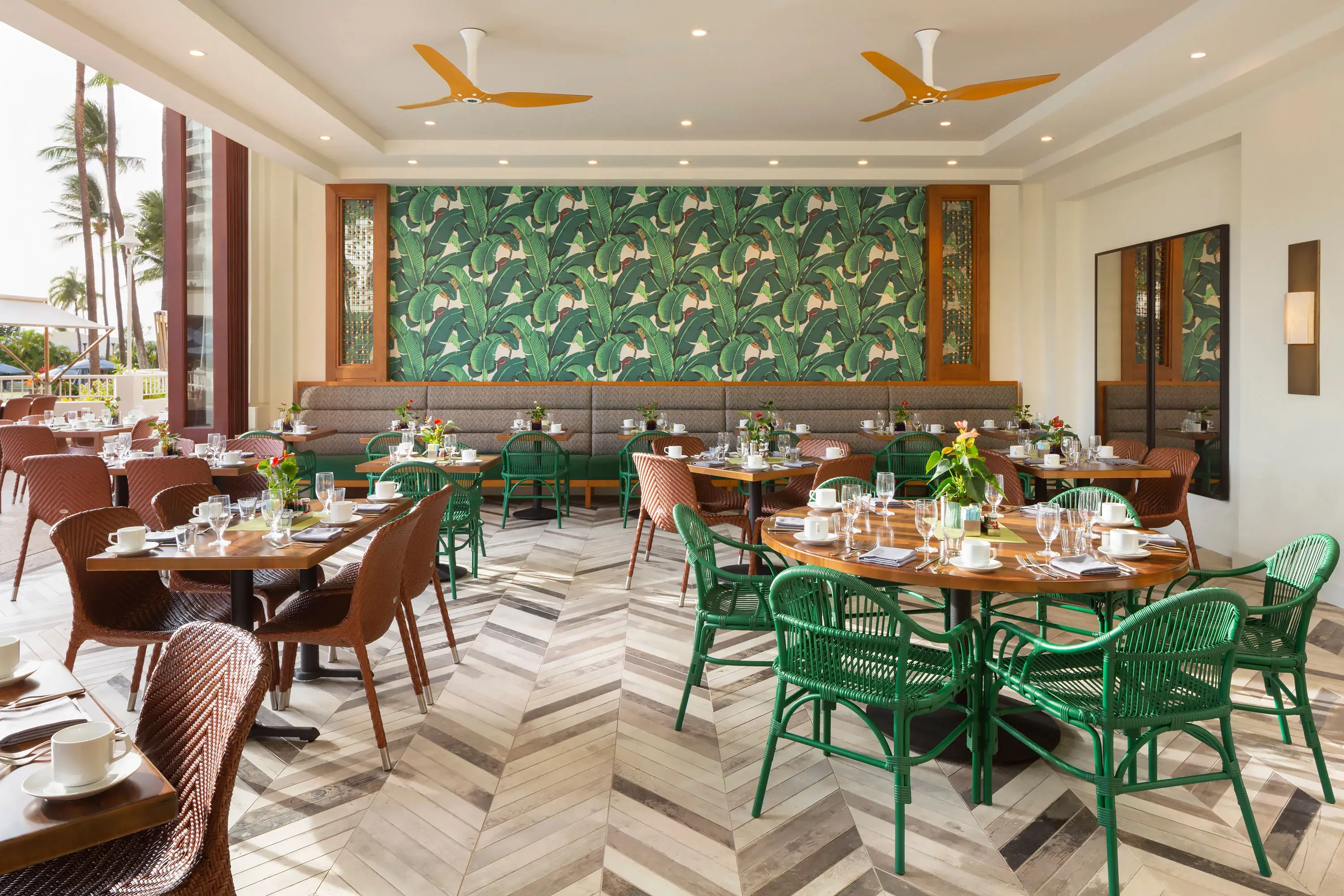 The airy interiors of Kea Lani Restaurant featuring brown and green tables and chairs on parquet flooring with a large patterned mural behind