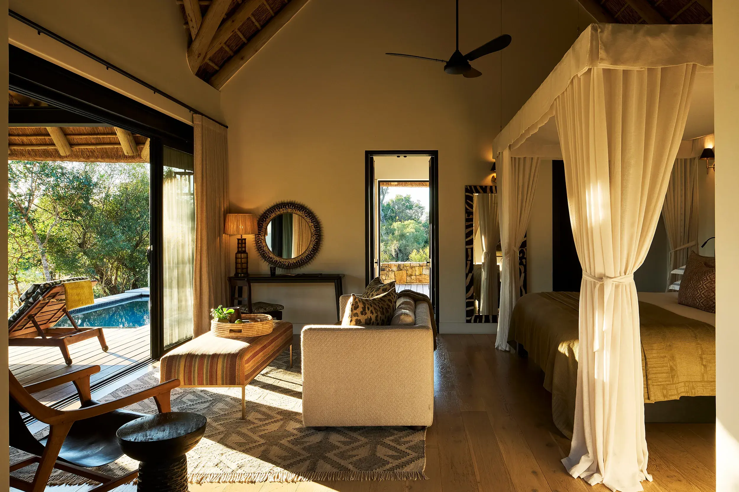 The interior of a suite at Monwana Game Reserve featuring a comfy lounge area in front of a four poster bed with white mesh curtains and a glimpse of a deck with a pool through large sliding doors