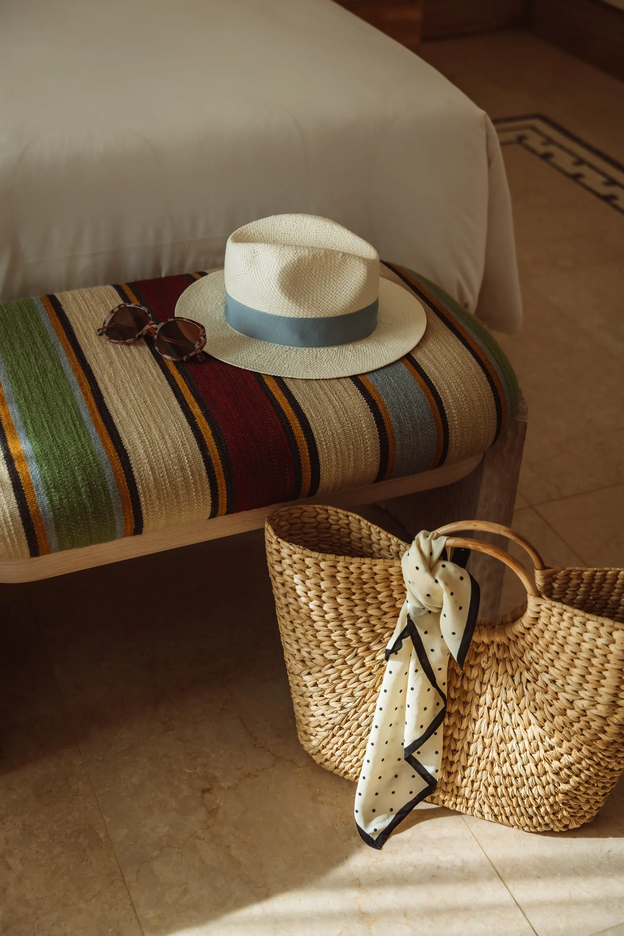 Straw hat with sunglasses on a striped bench beside a woven basket bag with a polka dot scarf