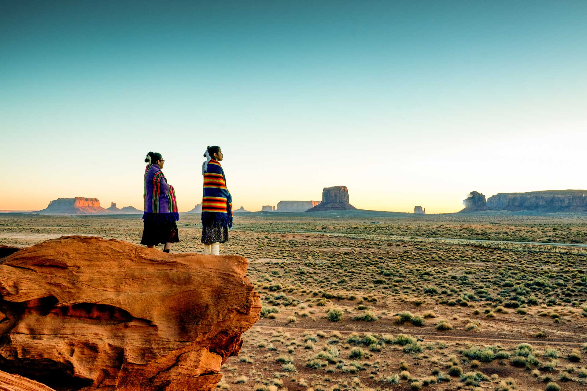 Two people standing on a rocky overlook wrapped in traditional blankets, gazing across a vast desert landscape.
