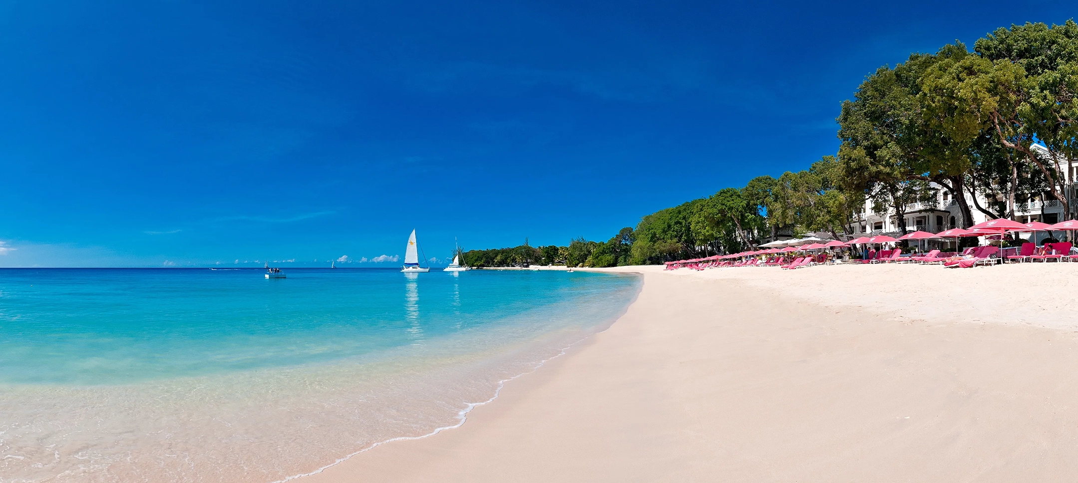 The beach at Sandy lane Barbados,with pink sun loungers facing the shoreline and two boats on the water