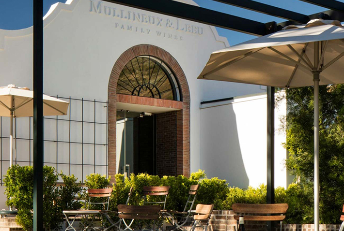 Outdoor seating area with wooden chairs and umbrellas in front of the Mullineux & Leeu Family Wines building, featuring a white facade and arched brick doorway.