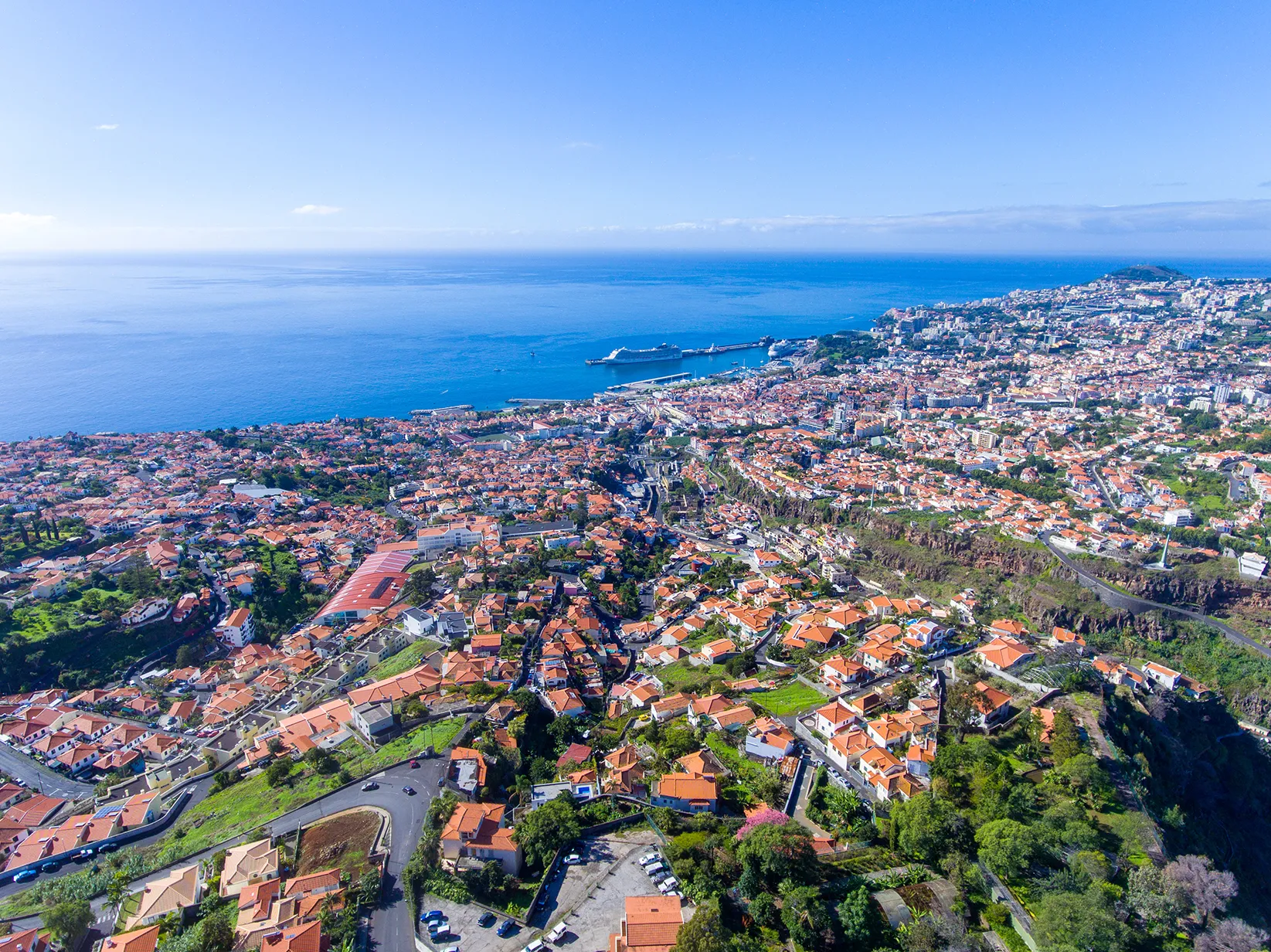 Aerial view of a coastal city with red-roofed buildings, a harbor, and the ocean stretching to the horizon.