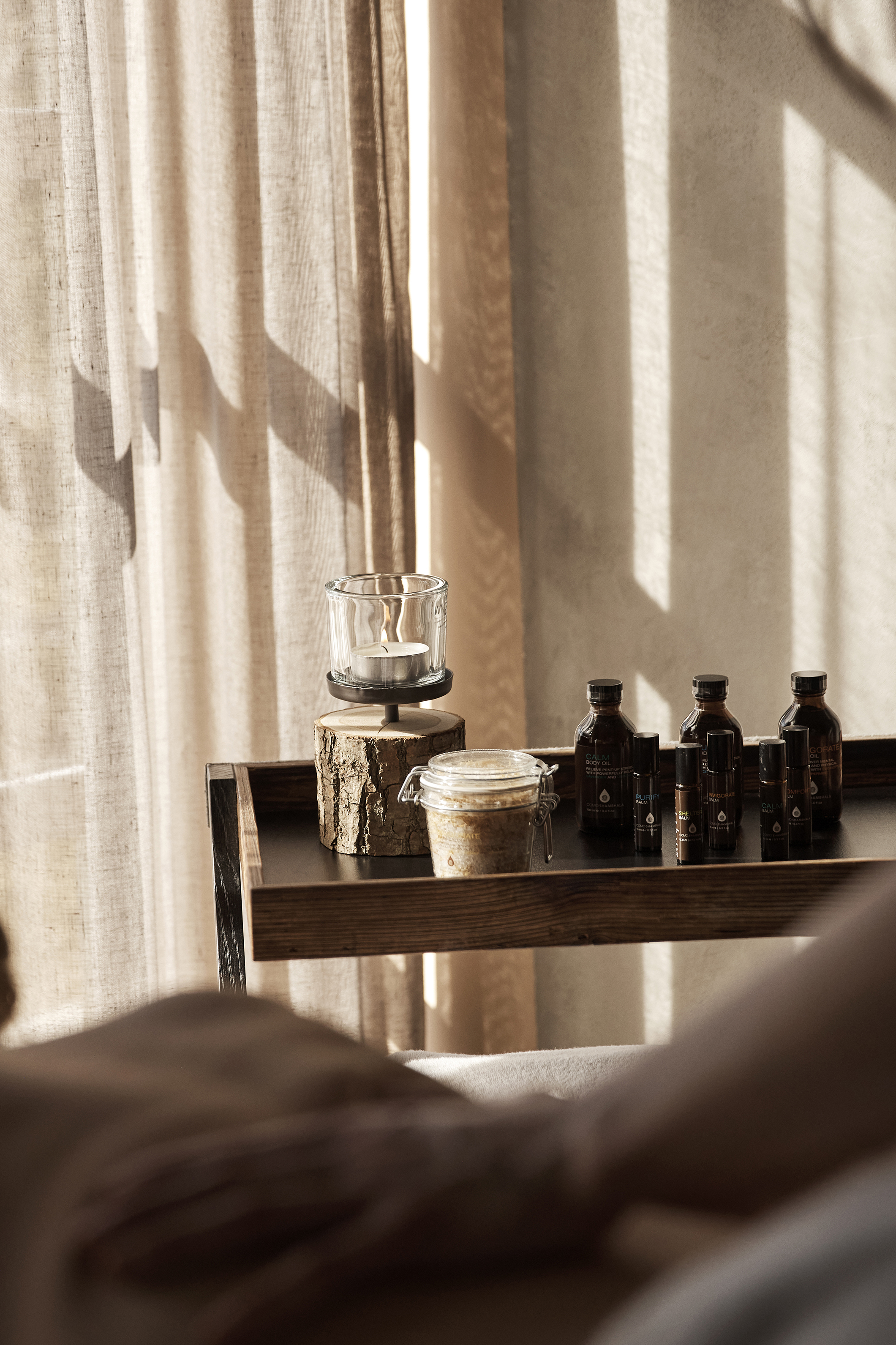A wooden shelf with various glass bottles, a candle and a jar against a backdrop of sheer curtains and soft sunlight, creating a serene and tranquil atmosphere.