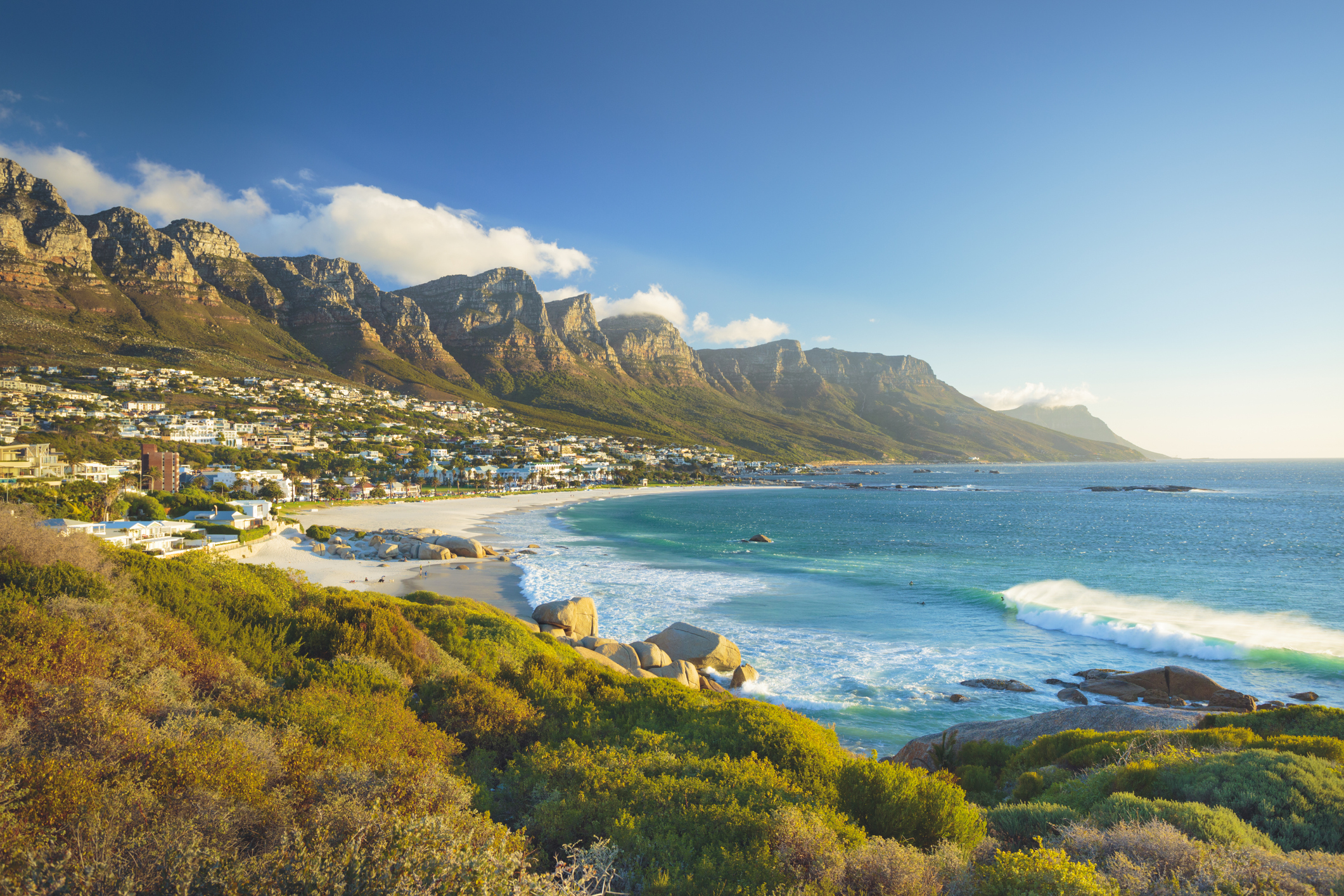 A scenic view of a coastal town with a sandy beach, turquoise sea, and a dramatic mountain range in the background under a clear blue sky.