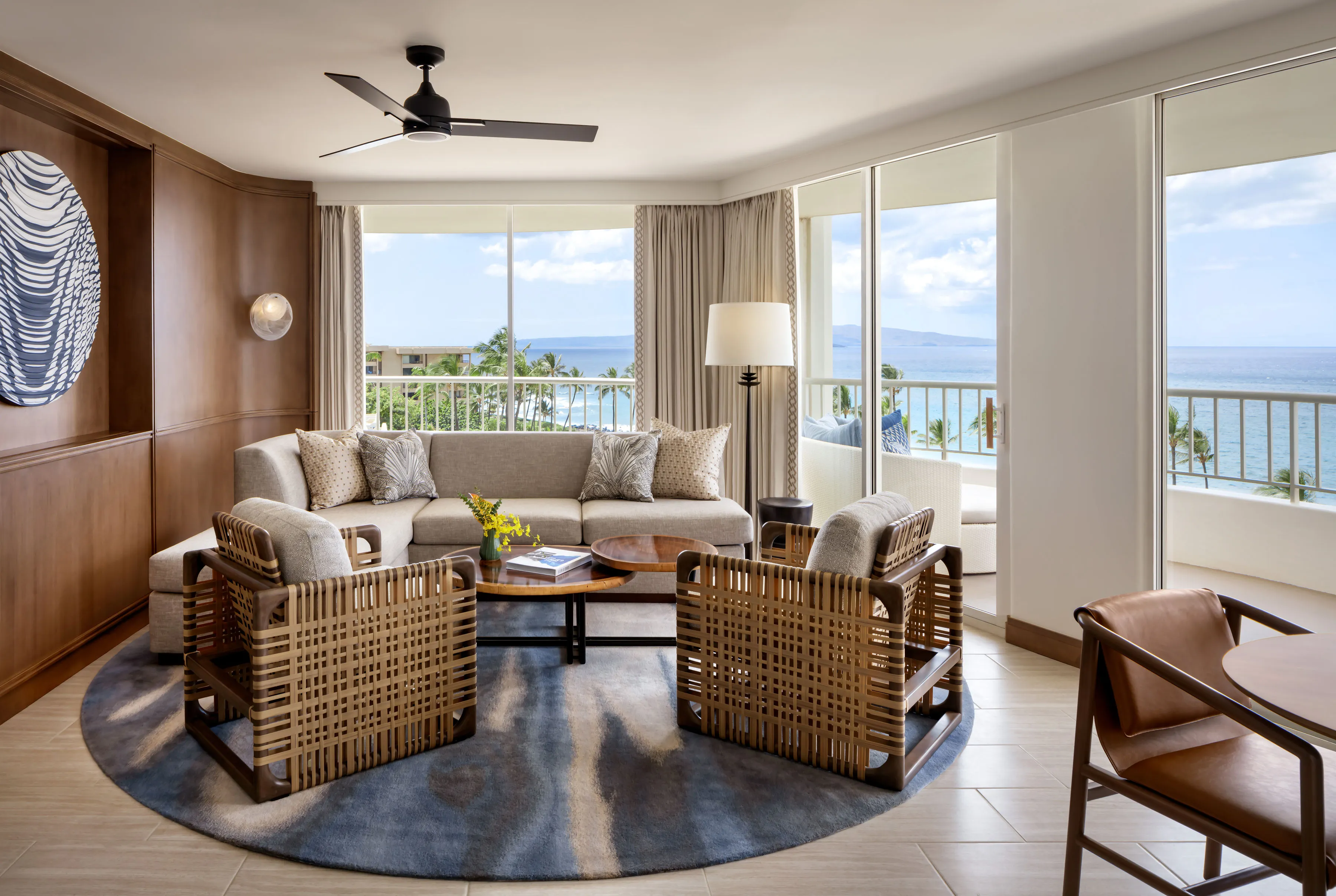 The comfy living room of a Corner Suite at Fairmont Kea Lani featuring a plush sofa, woven single chairs, blue furnishings and semi wrap around windows