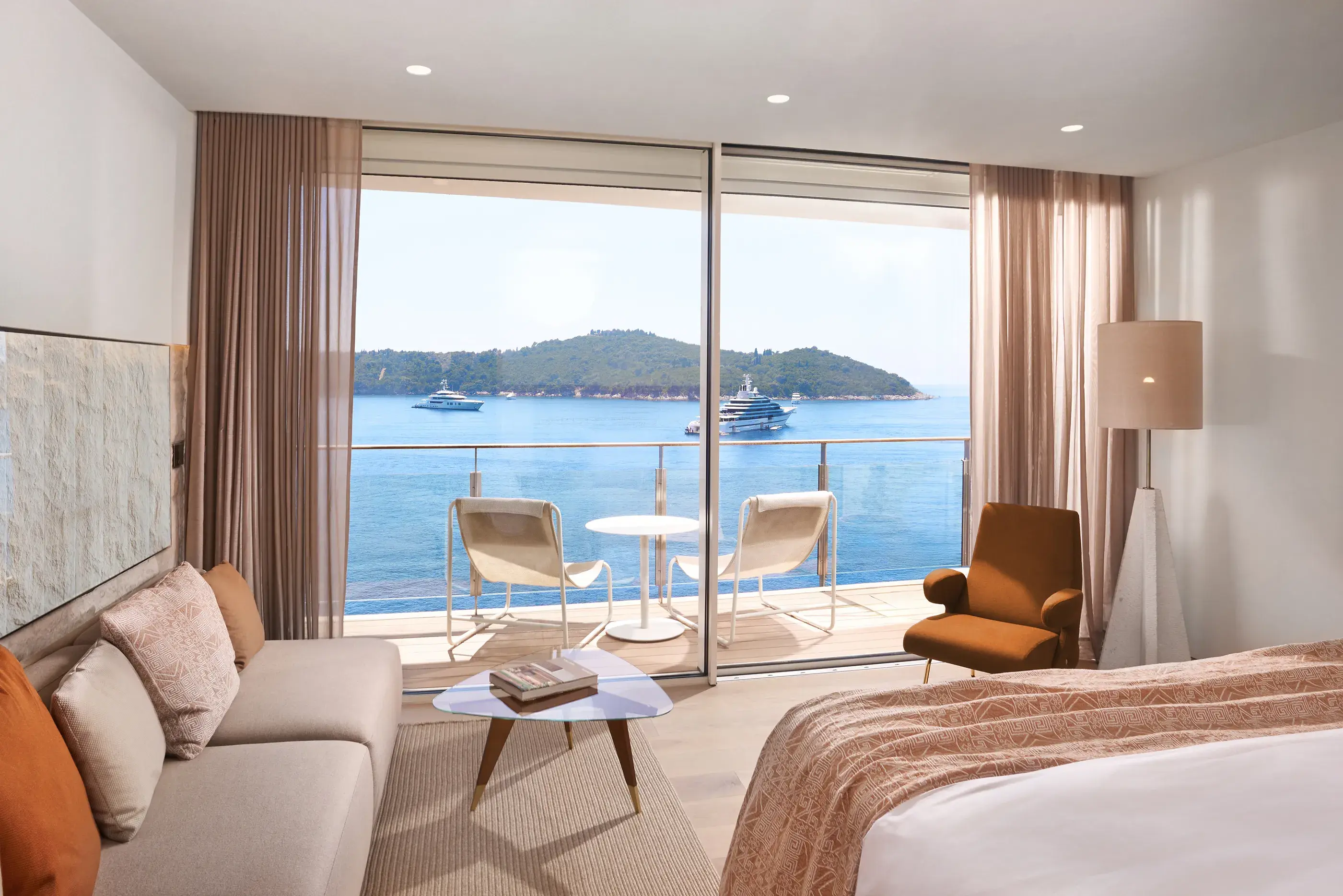 Deluxe Grand Sea View room at Villa Dubrovnik in Croatia featuring a stylish lounge area with warm toned furniture and large balcony windows looking out to sea with ships and Lokrum Island