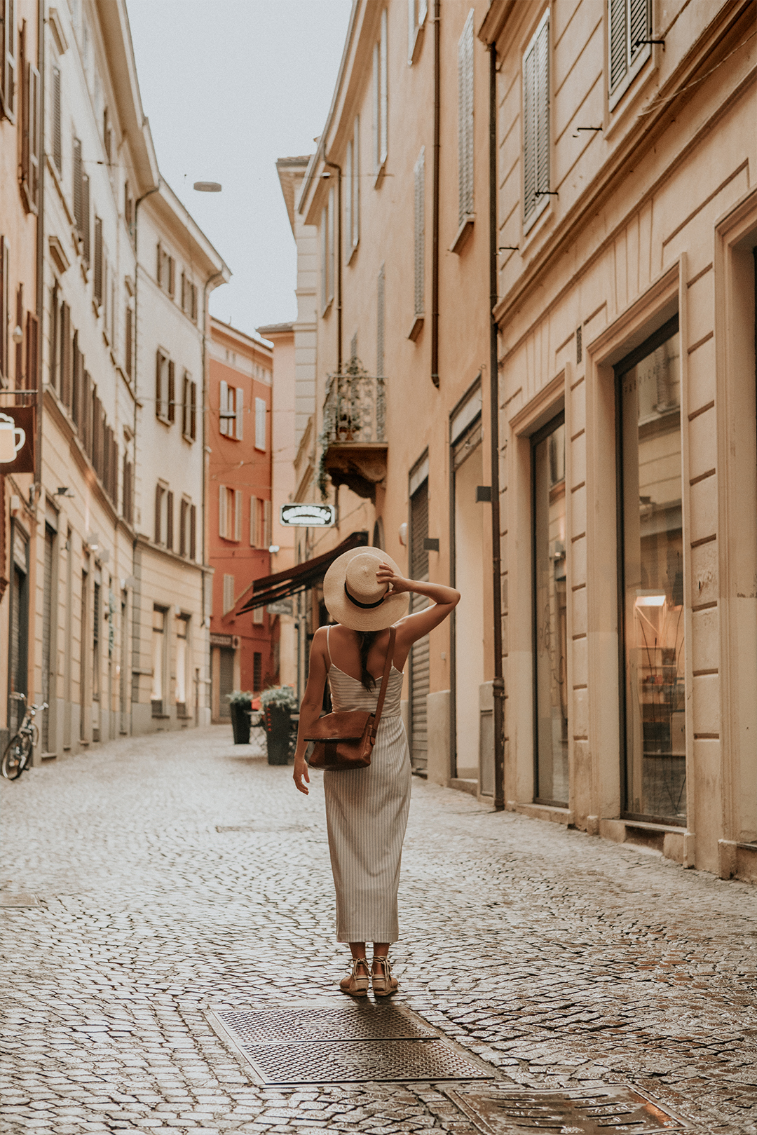 A person stands in the centre of a cobblestone street in an old European city, facing away from the camera, wearing a hat and a striped dress.