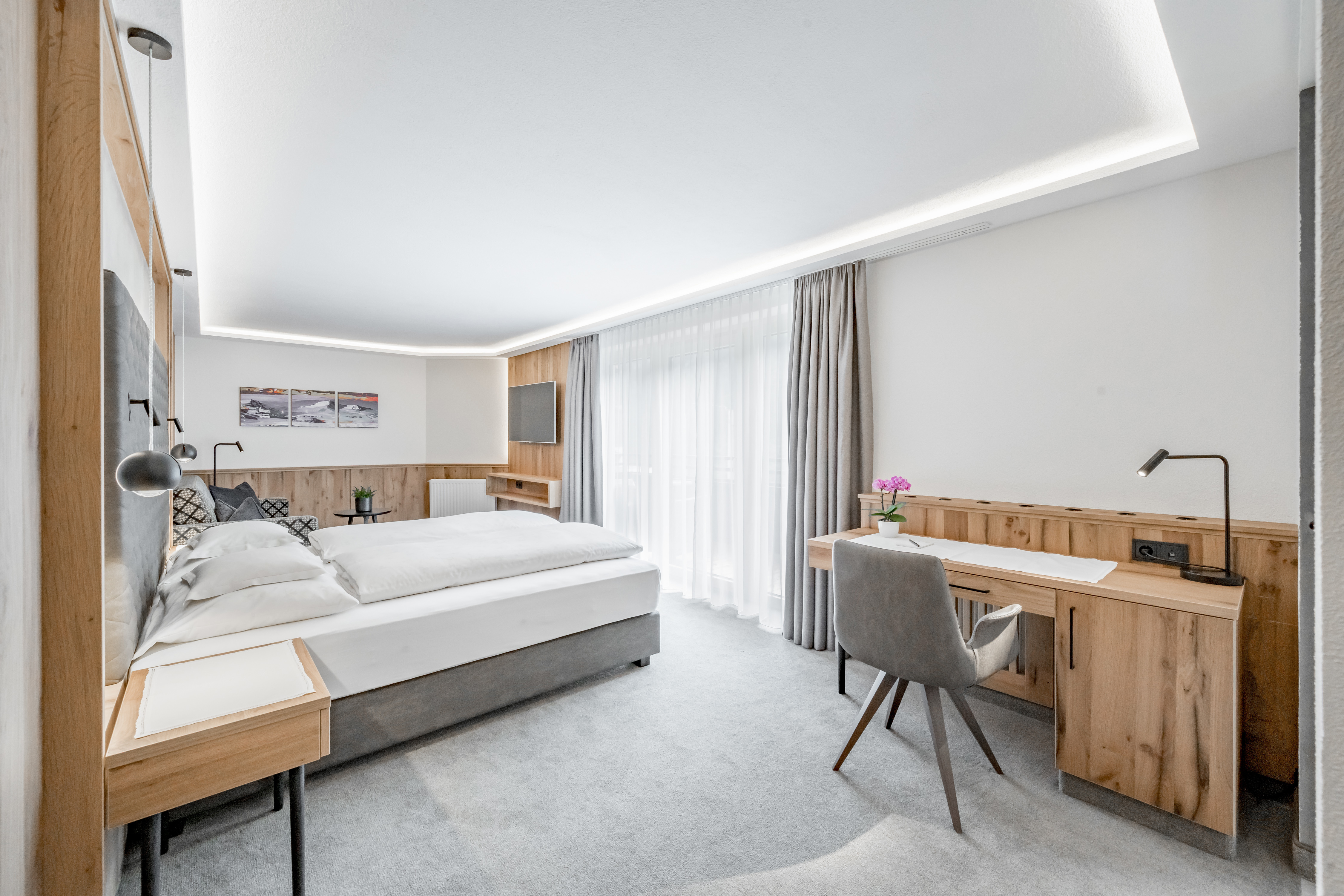 A modern hotel room with a large bed, wooden furniture, a desk with a chair, and a window with sheer curtains allowing natural light to fill the space