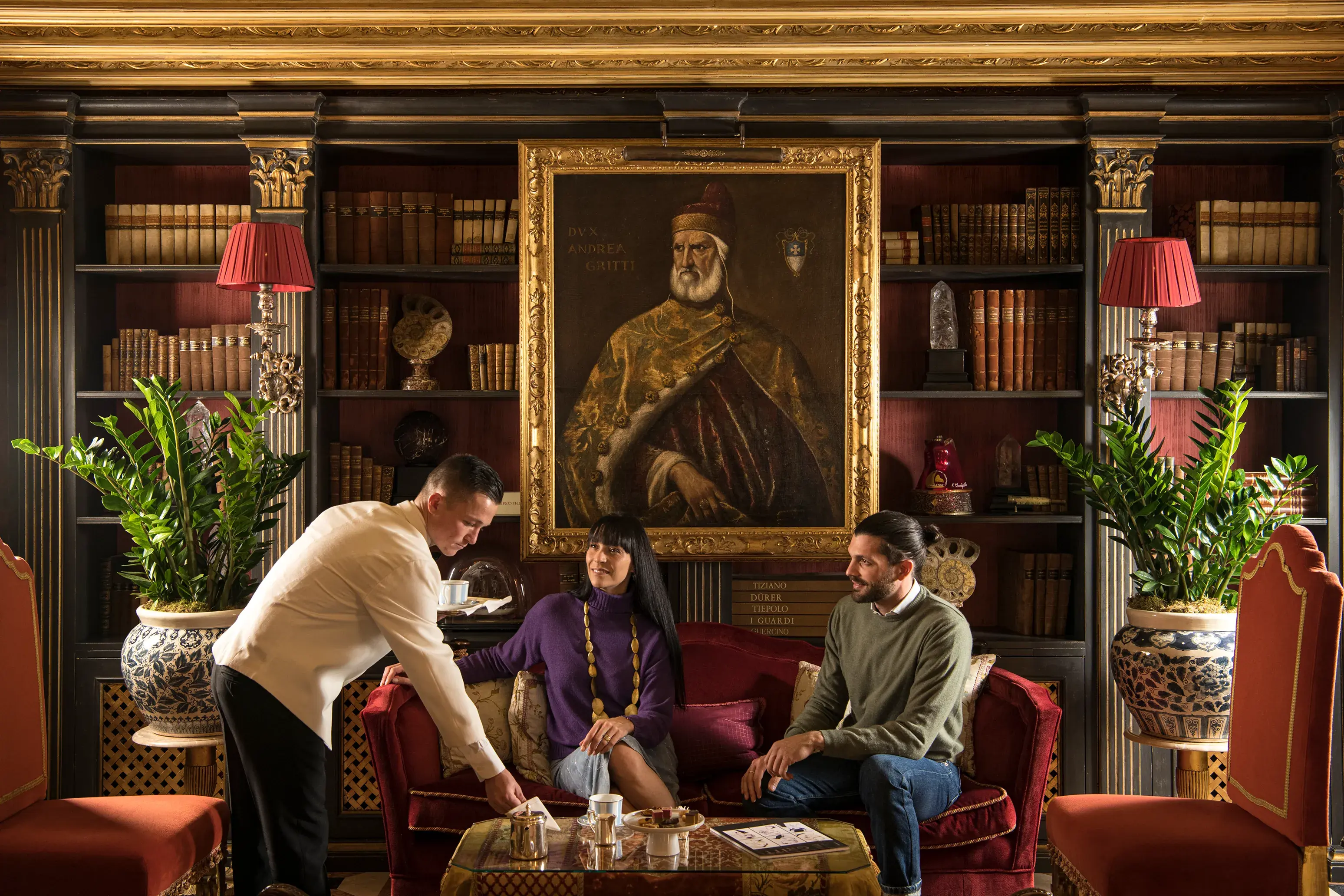 Luxurious library lounge at The Gritti Palace in Venice featuring antique bookshelves, ornate gold-framed portrait, and elegant seating.