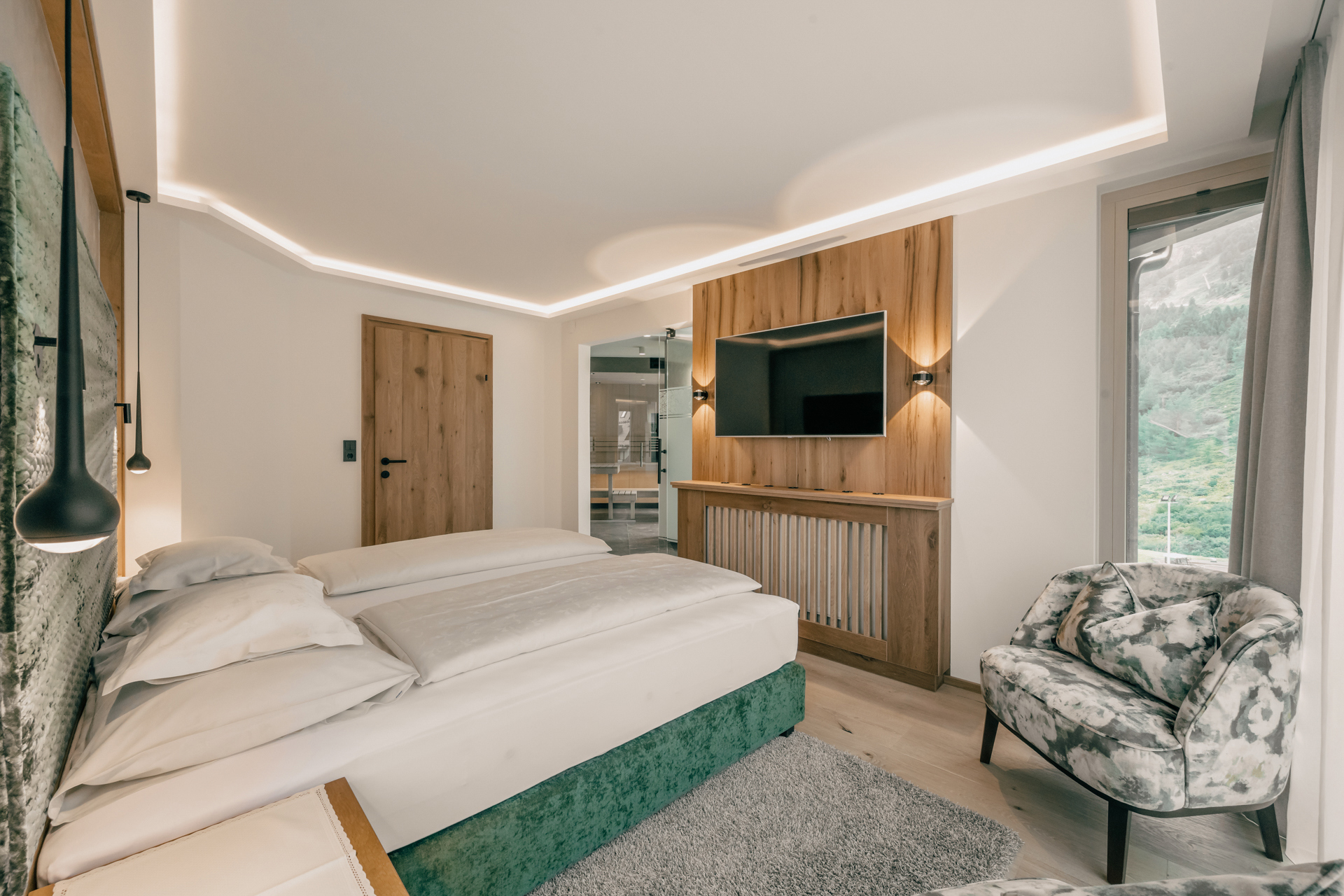 A modern Spa Suite at the Gourmet and Wine Hotel Austria featuring a large bed, wooden accents, a wall-mounted TV, and a patterned armchair by the window