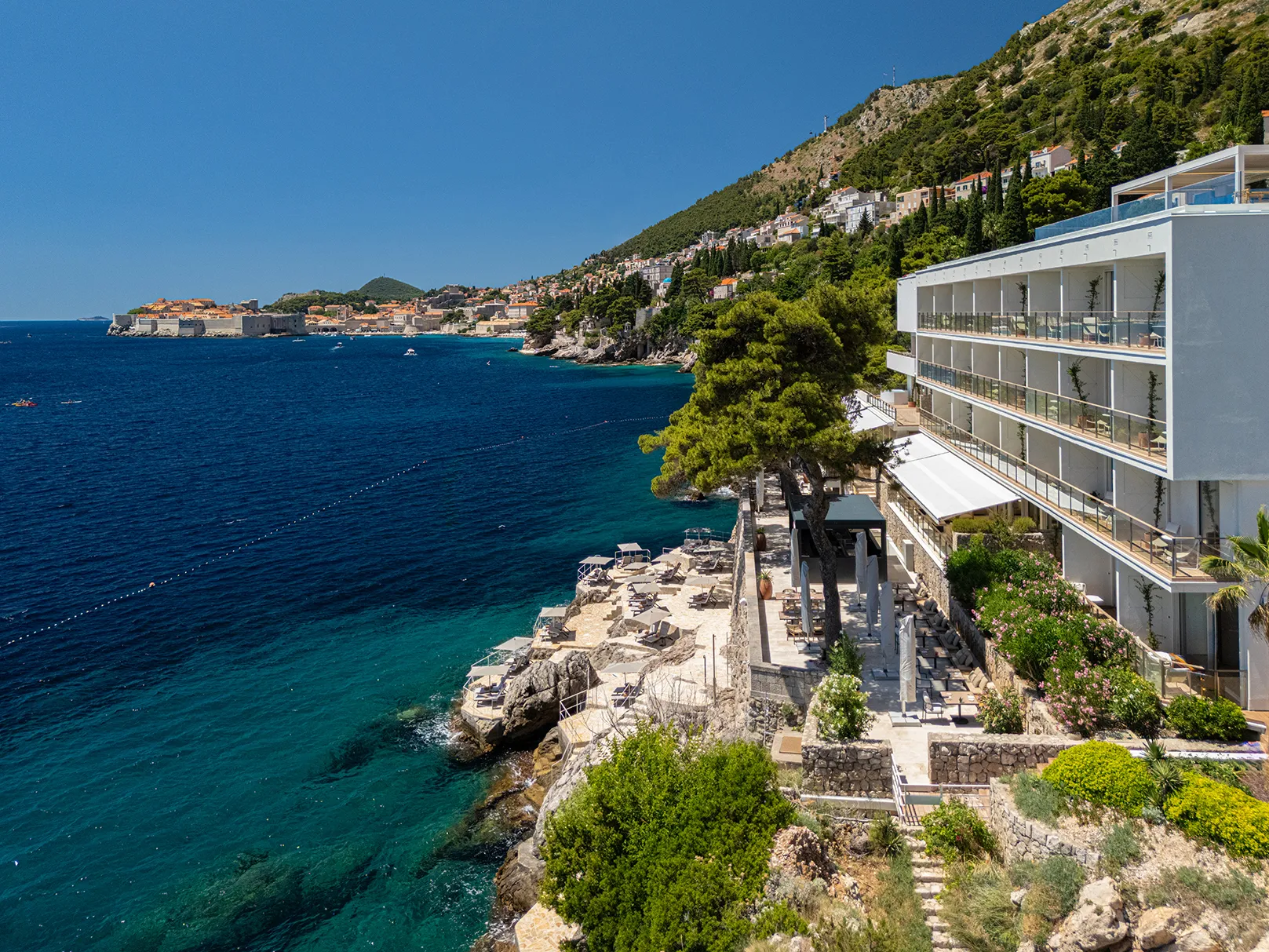 Clifftop seaside hotel overlooking turquoise waters and a historic coastal city.