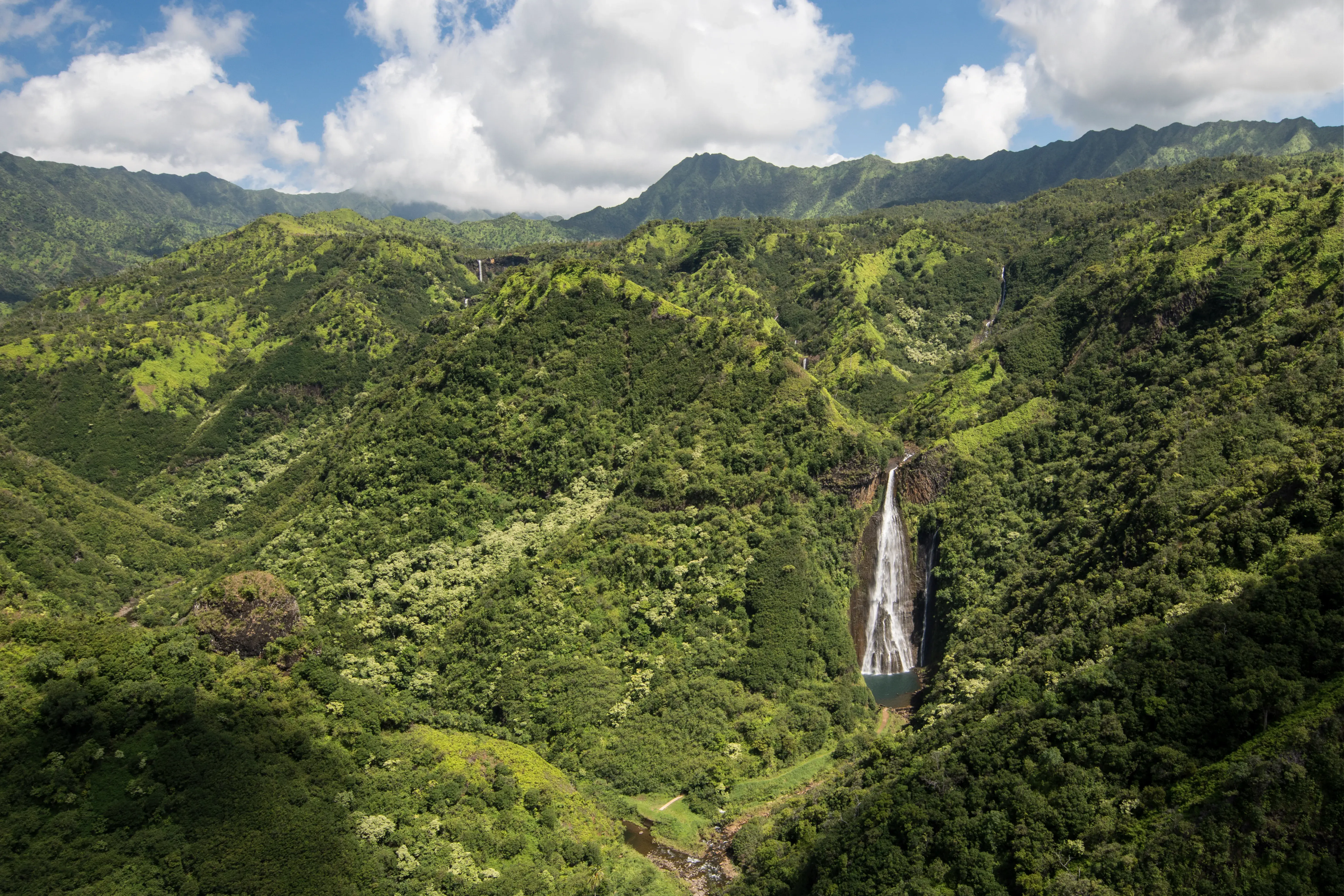 A shot of Mānāwaiopuna Falls from across the valley featuirng the waterfall surrounded by green hills beneath a bright sky