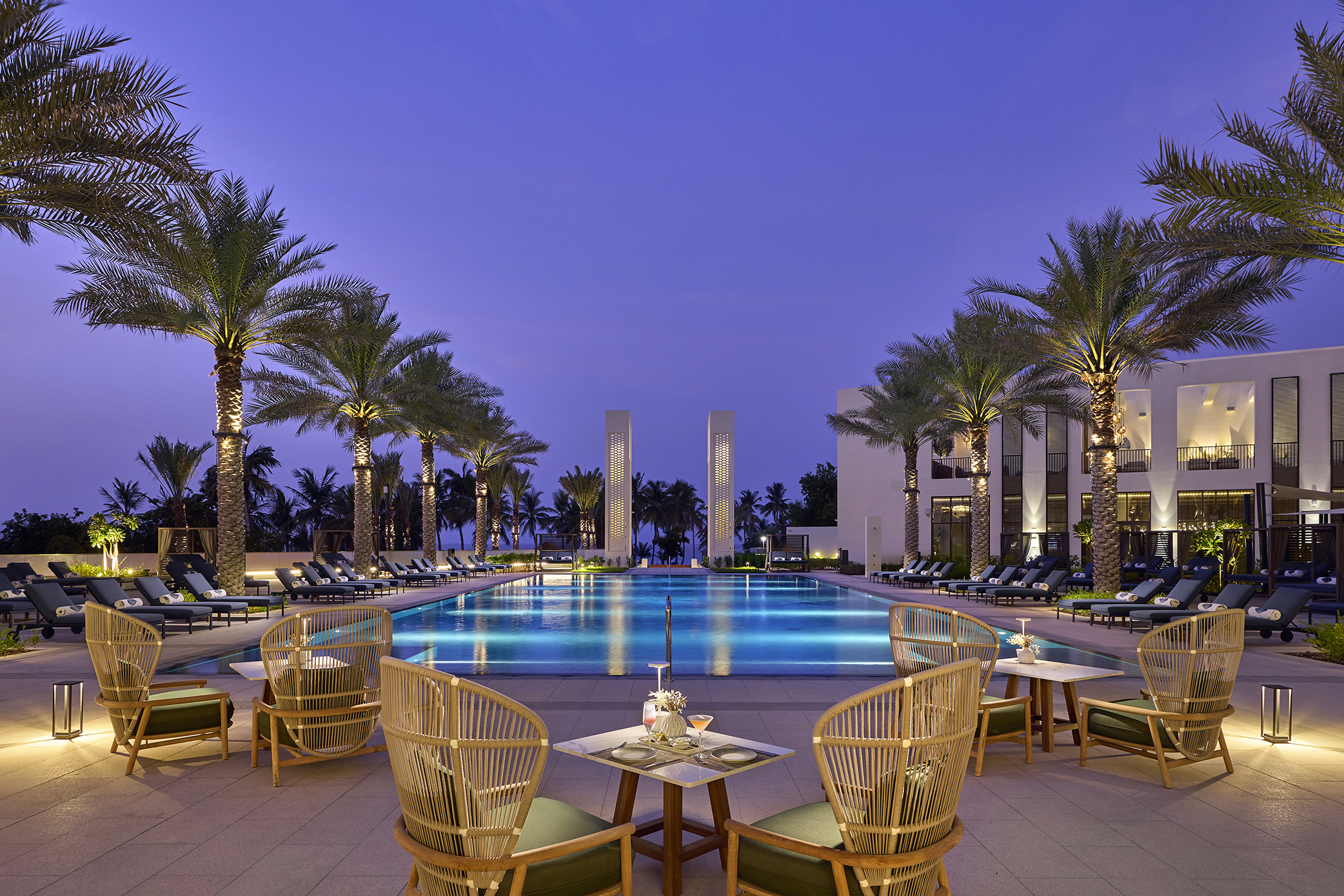 The atmospheric pool of Mandarin Oriental Muscat lit up at night and surrounded by rattan pool furniture and dining tables