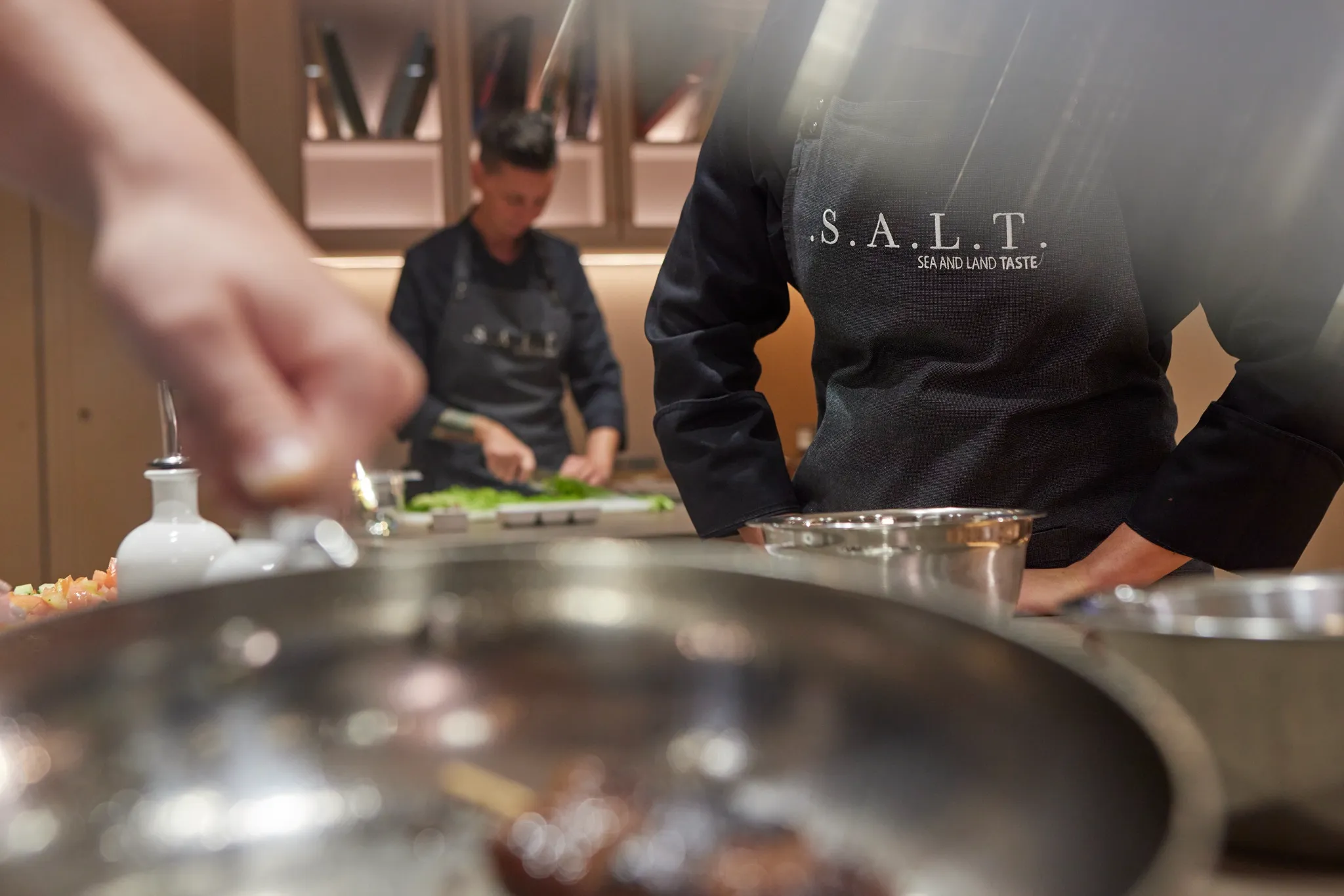 Chefs in "S.A.L.T. Sea and Land Taste" aprons preparing food in a kitchen, with one chopping vegetables and another near a pan.