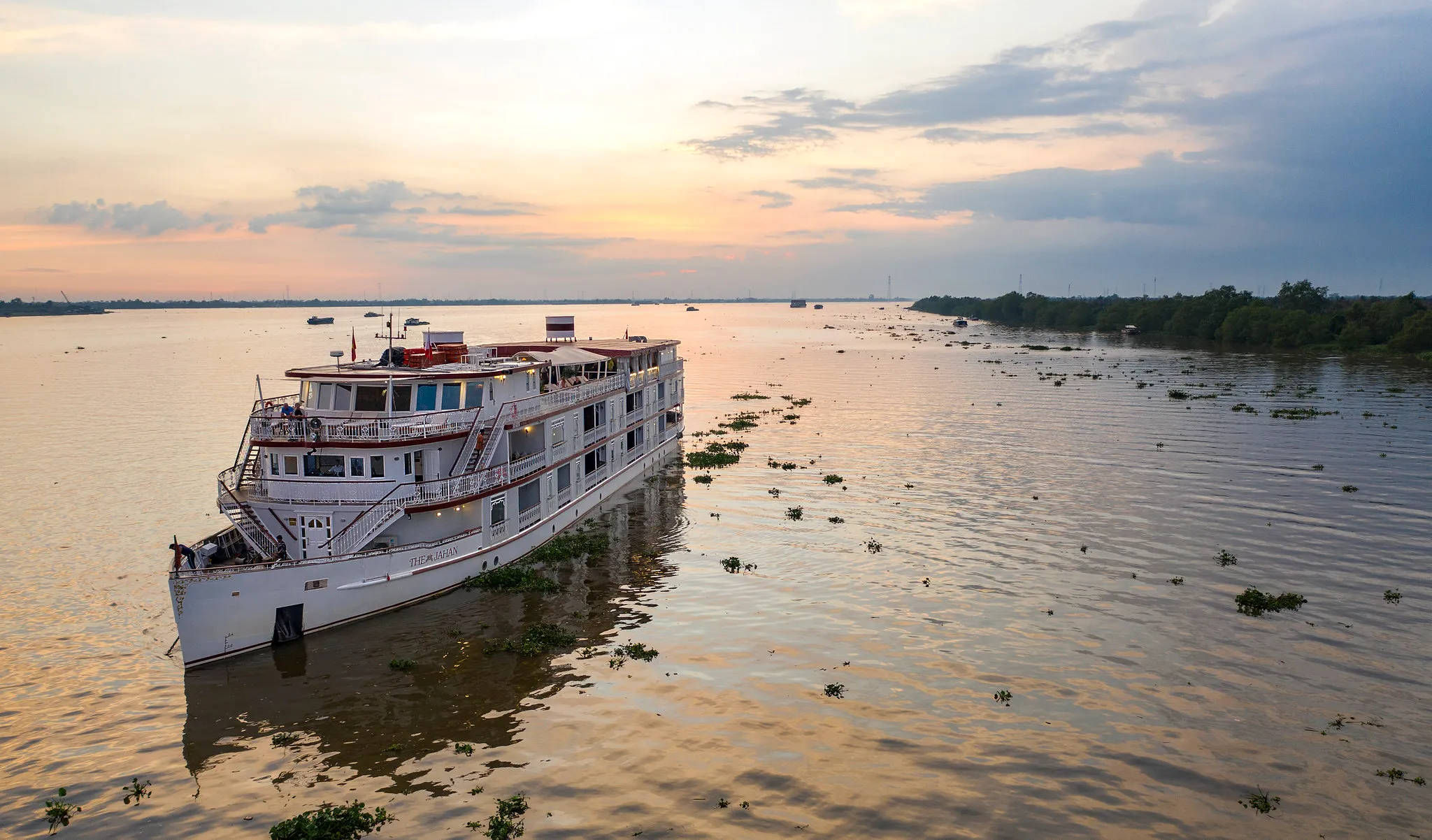 A large white Heritage Line riverboat cruises along a calm river at sunset, with floating vegetation and a tree-lined shoreline in the distance
