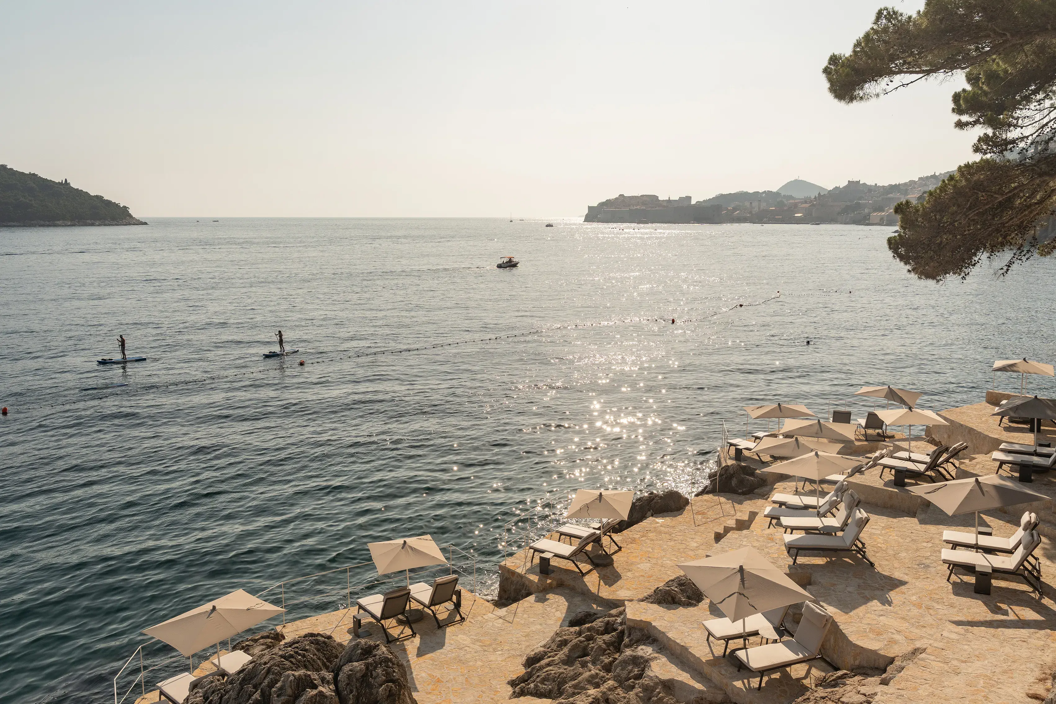 Beach area at Villa Dubrovnik in Croatia featuring sun loungers on a stone terrace beside the water with two people paddle boarding 