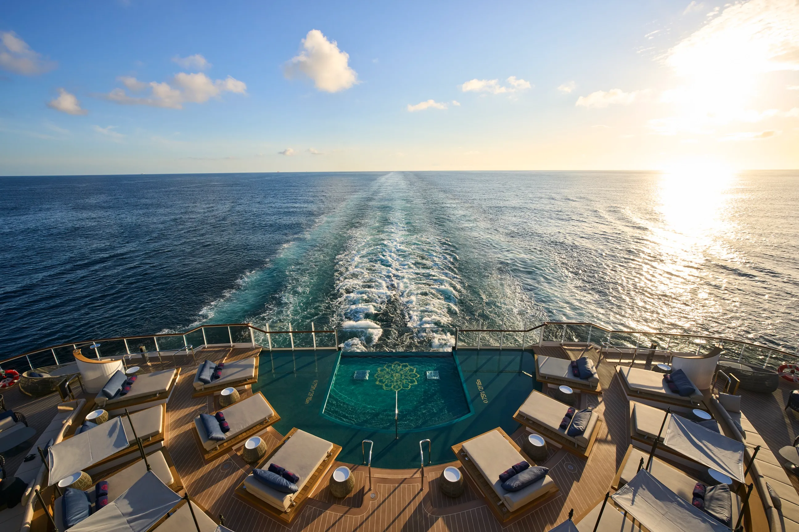 Elegant cruise ship deck featuring a tranquil pool and sun loungers, with ocean views and a wake trailing into the horizon.