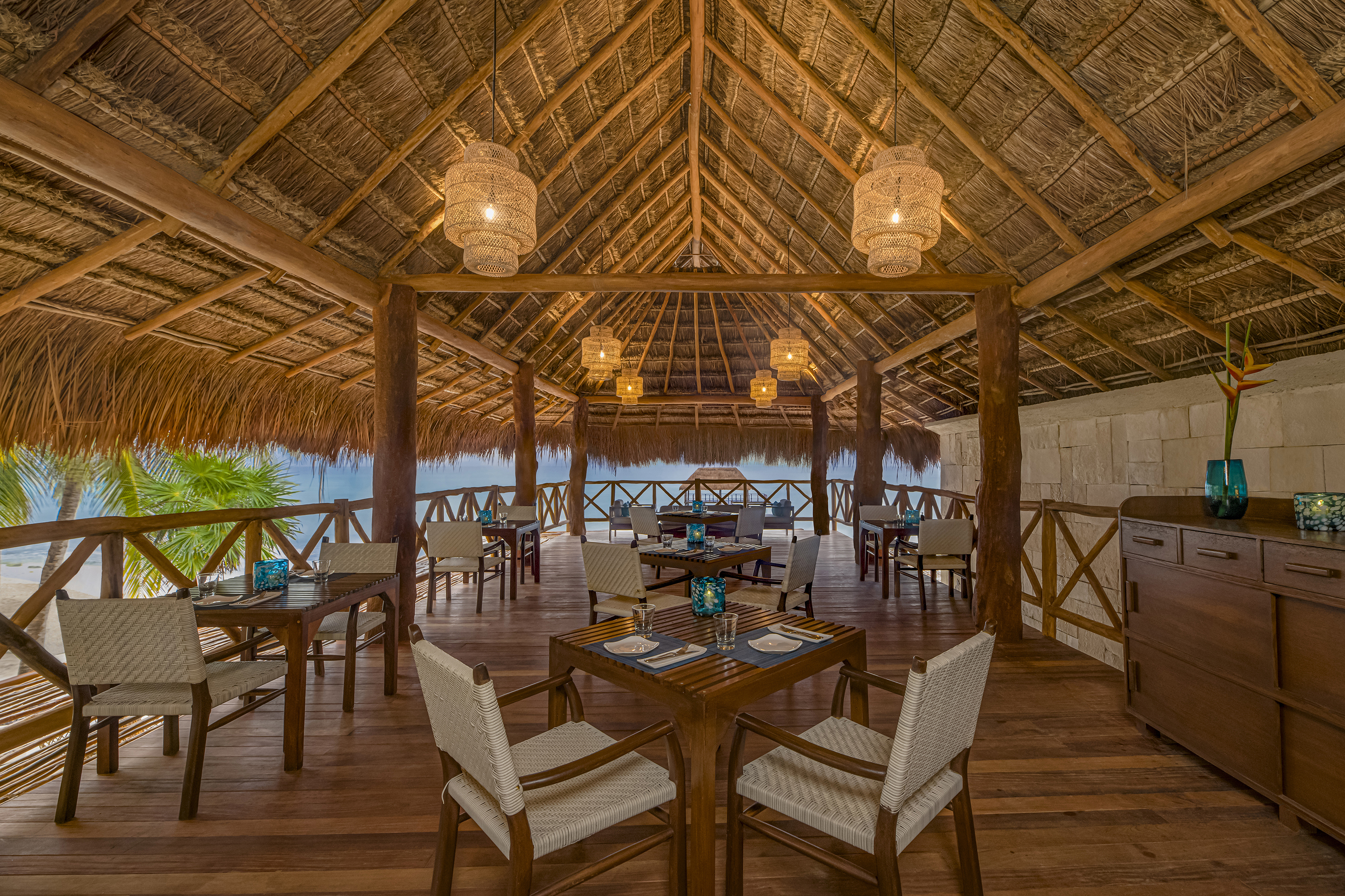 Several dining tables set for a meal beneath a vaulted thatched ceiling with rattan light shades at Coral Grill at Viceroy Riviera Maya