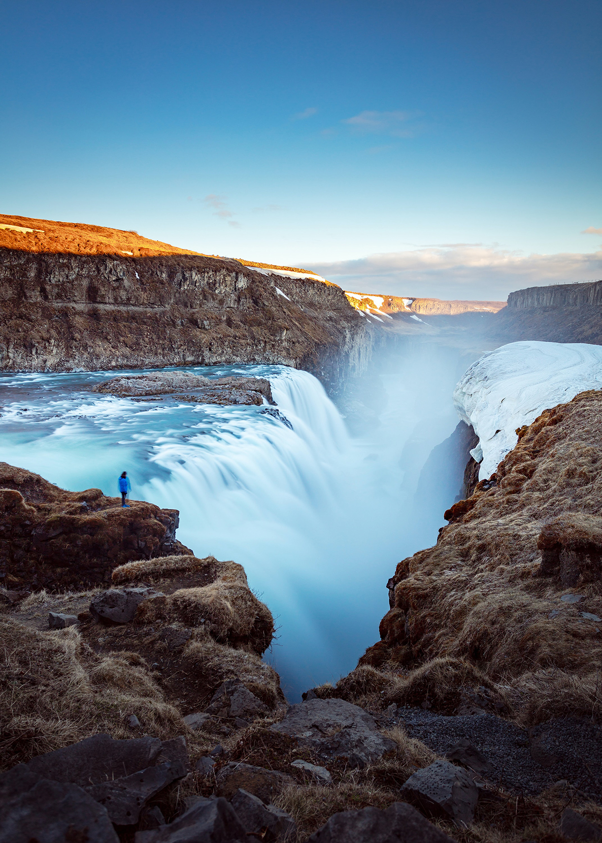 A large waterfall dropping into a crevasse, and a person looking from a rocky ledge