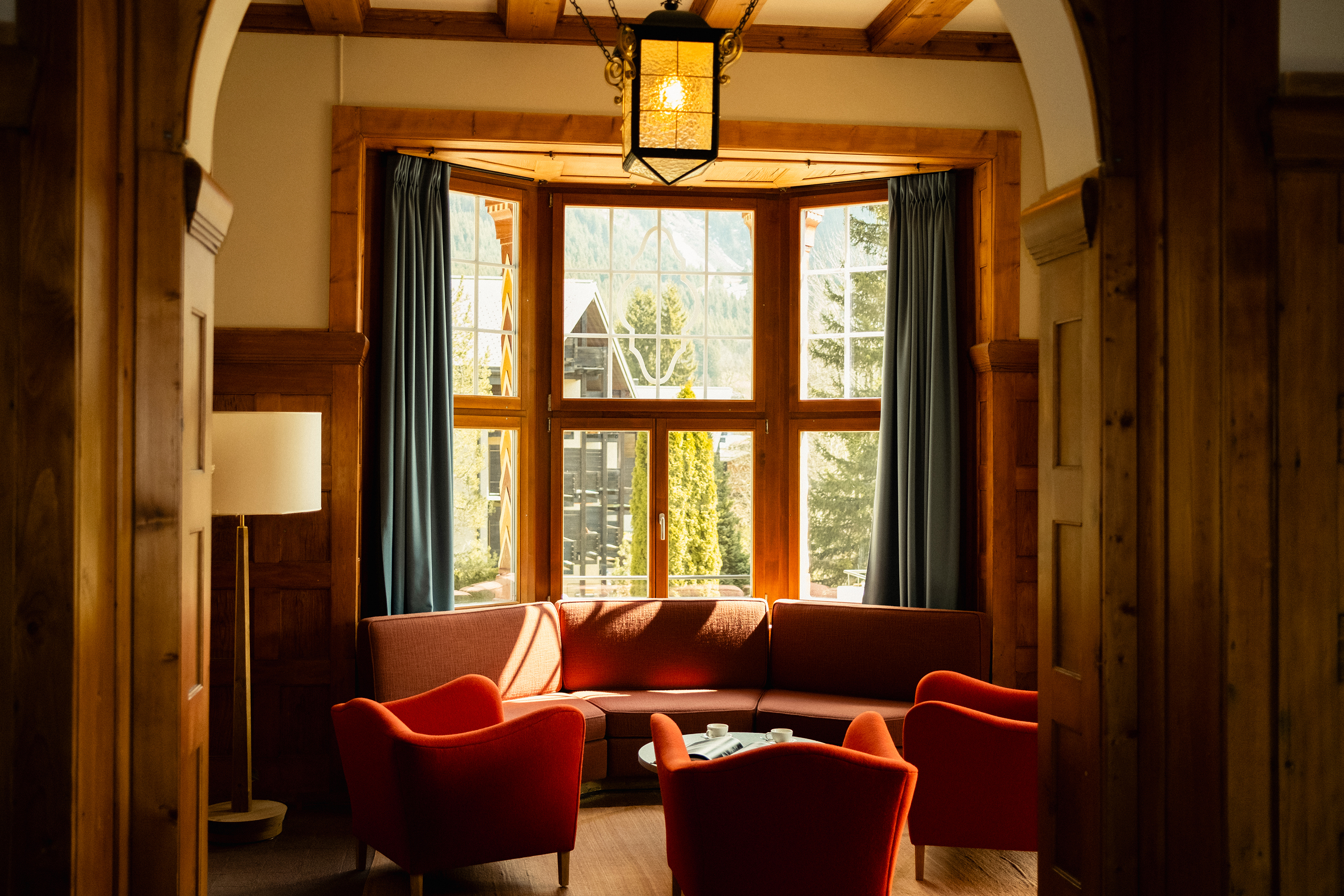 A cosy lounge area featuring red furniture in a wood panelled room in front of a bay window with an alpine scene outside