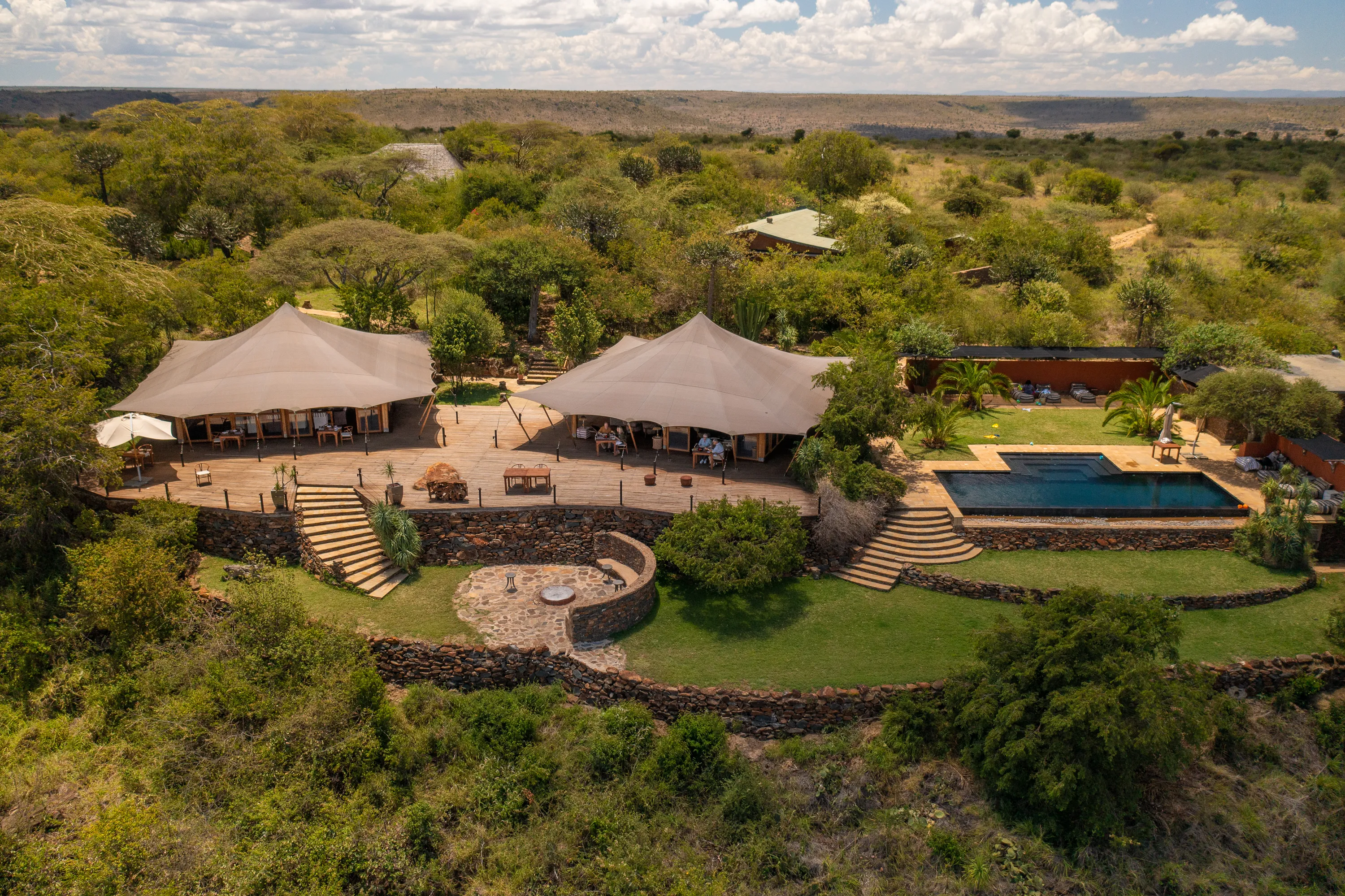 Aerial view of Elewana Loisaba Tented Camp featuring two main tented areas, a large deck, infinity pool and firepit surrounded by vegetation with a glimpse of other buildings behind