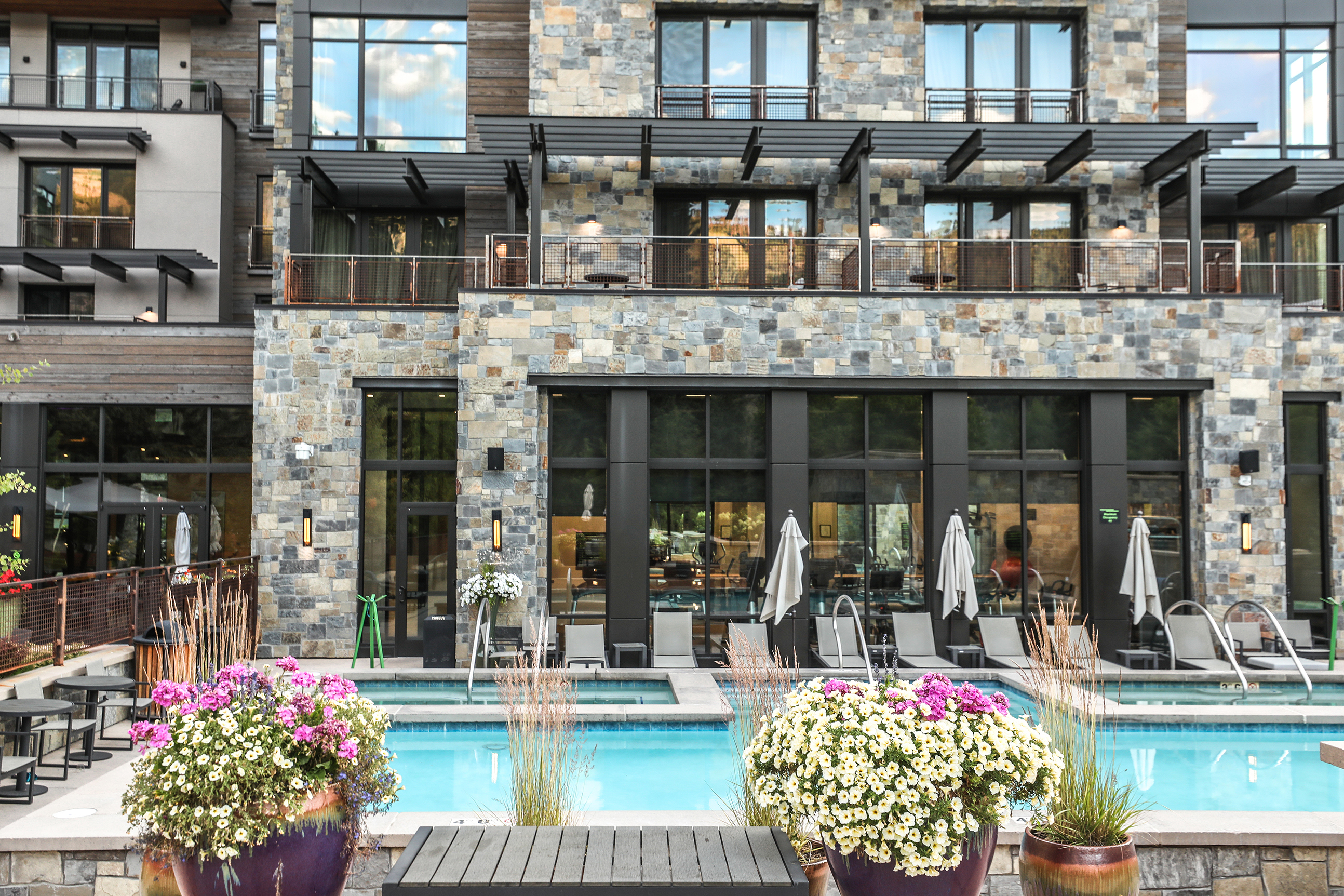 A modern stone building of Limelight Ketchum with large windows and balconies overlooking a swimming pool surrounded by lounge chairs and umbrellas, with colorful flower pots in the foreground